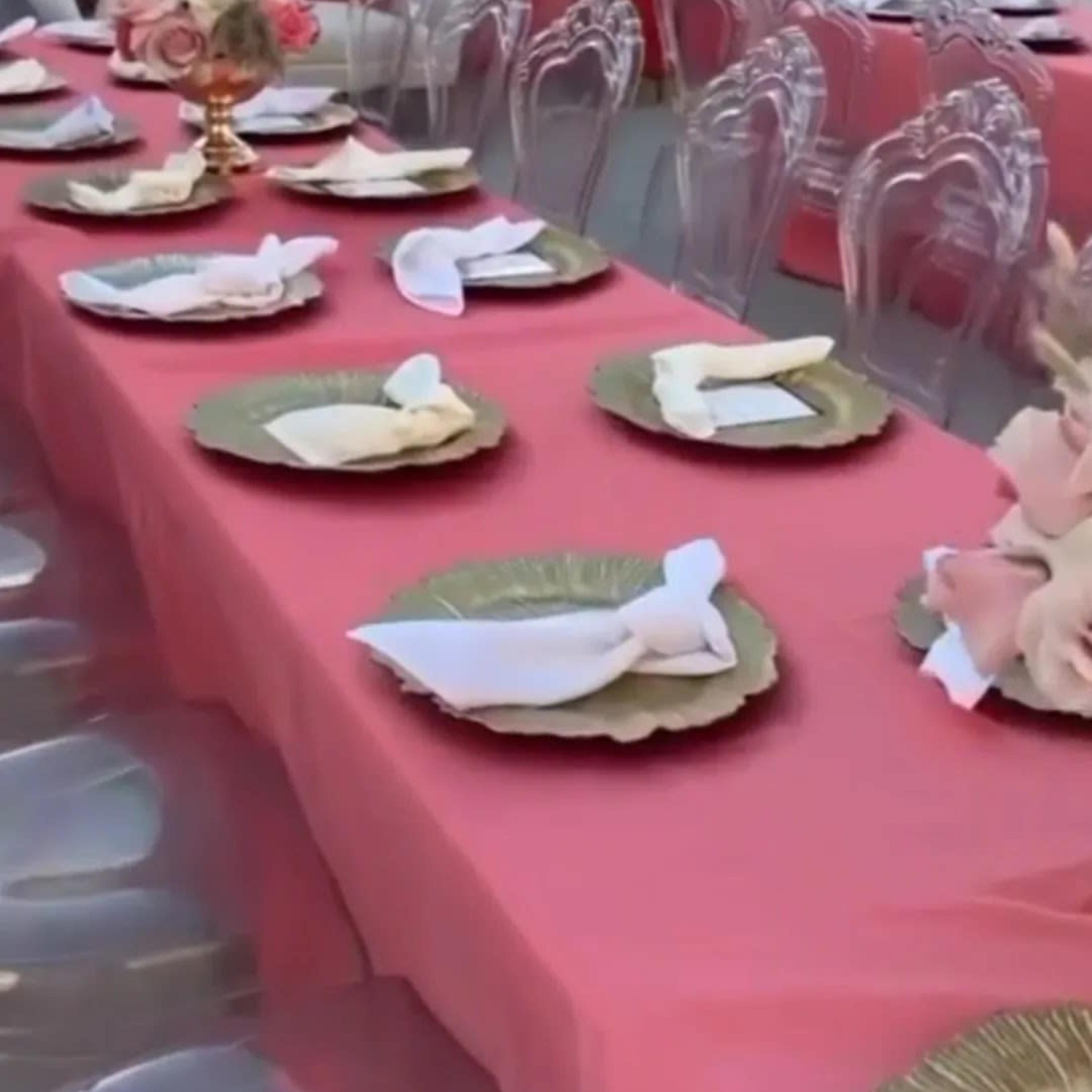 A long table is set with gold-rimmed plates and neatly folded napkins in an outdoor setting decorated with pink drapery and floral accents.
