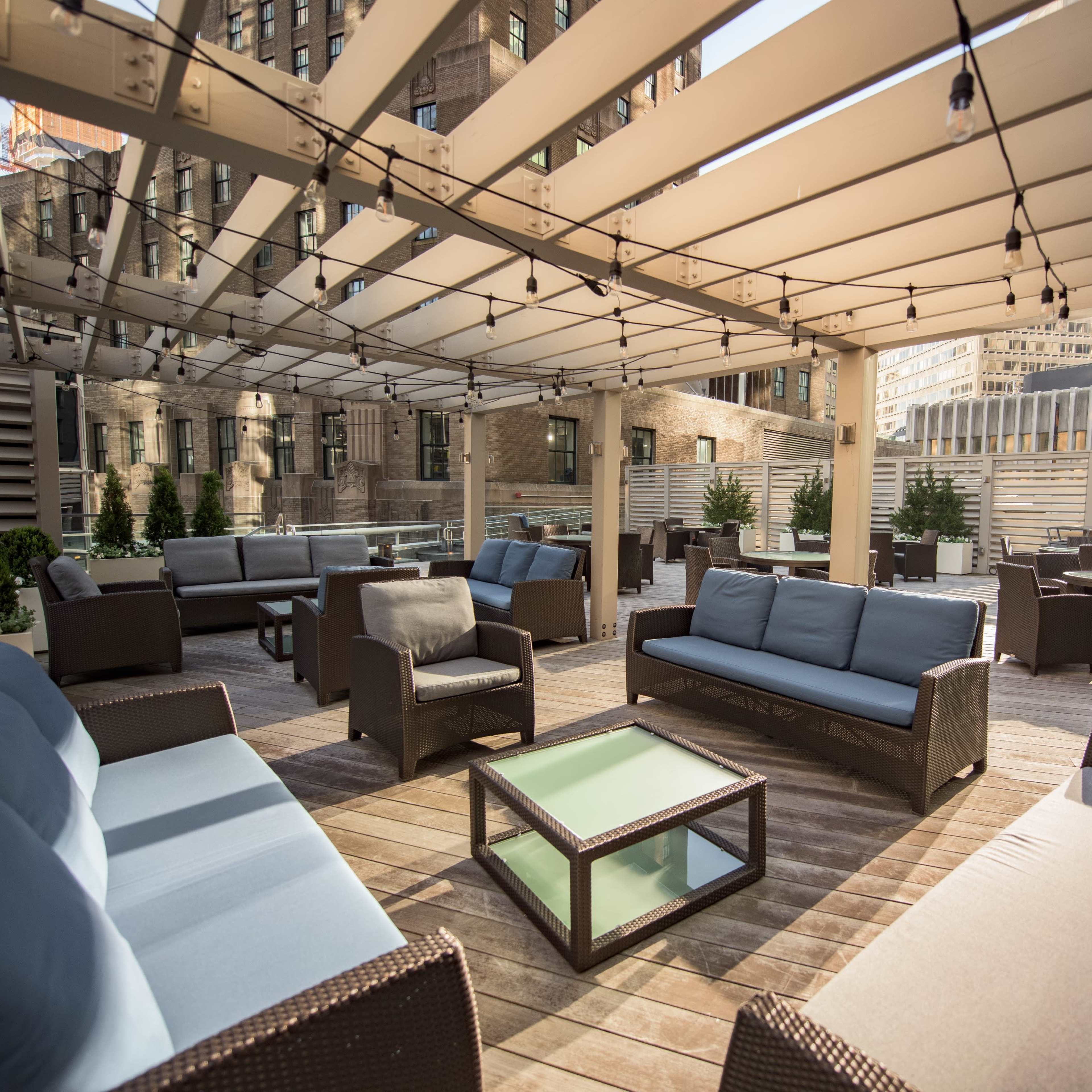 A rooftop patio with several seating areas, including sofas and tables, illuminated by string lights against a backdrop of urban buildings.