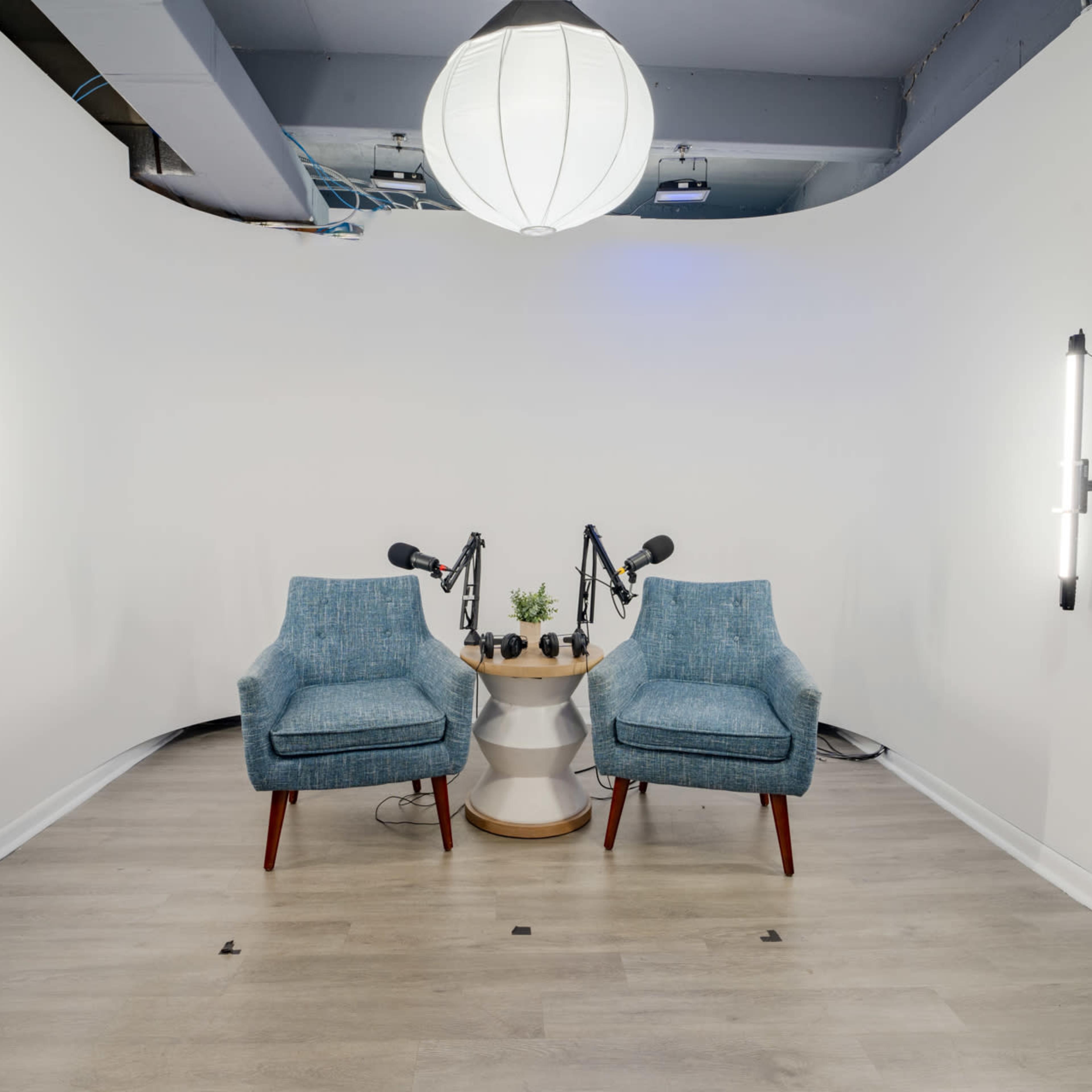 A minimalist recording studio with two blue chairs facing each other, a small round table between them, and microphones on stands attached to the walls.