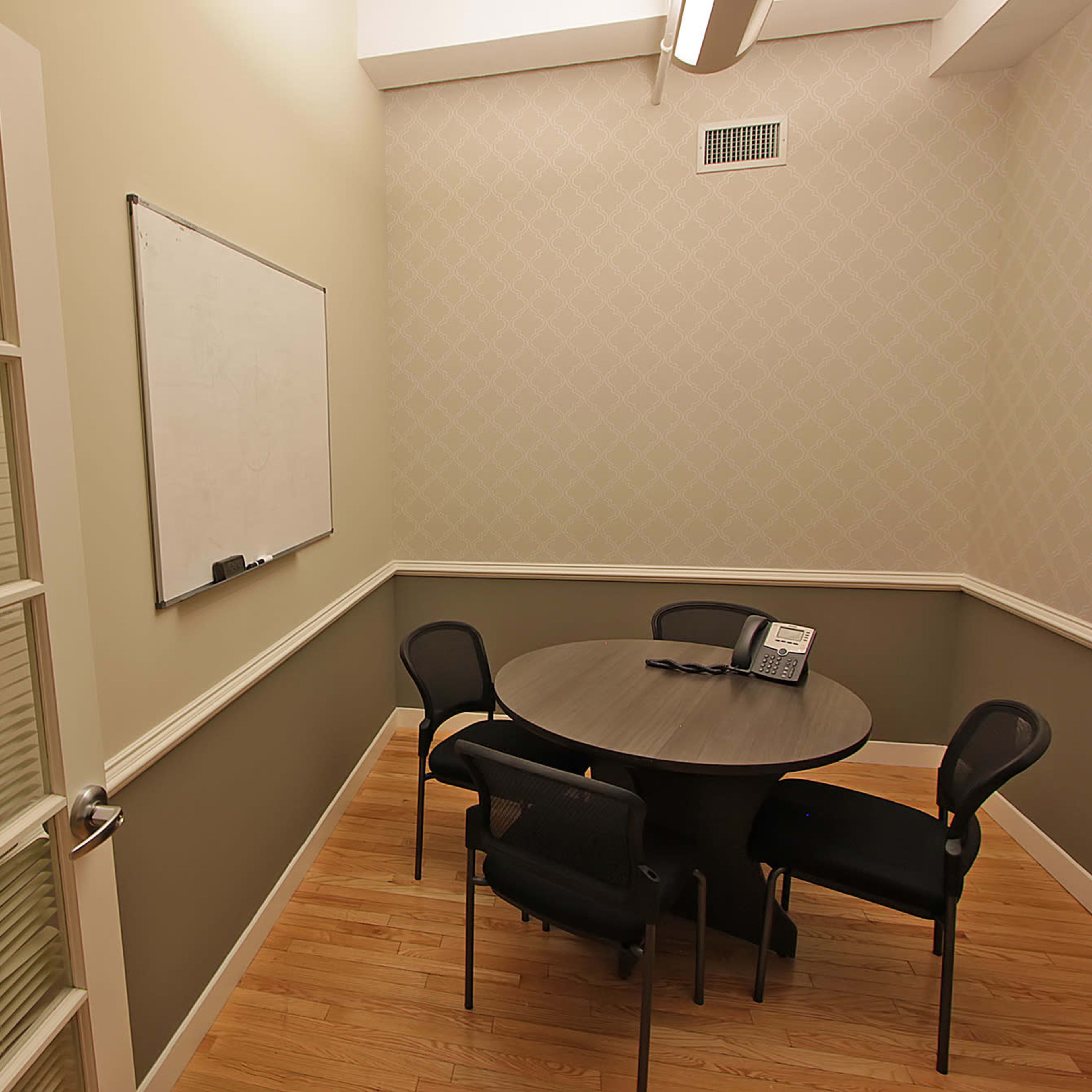 A small meeting room with a round table and four chairs, a whiteboard on the wall, and a telephone on the table.