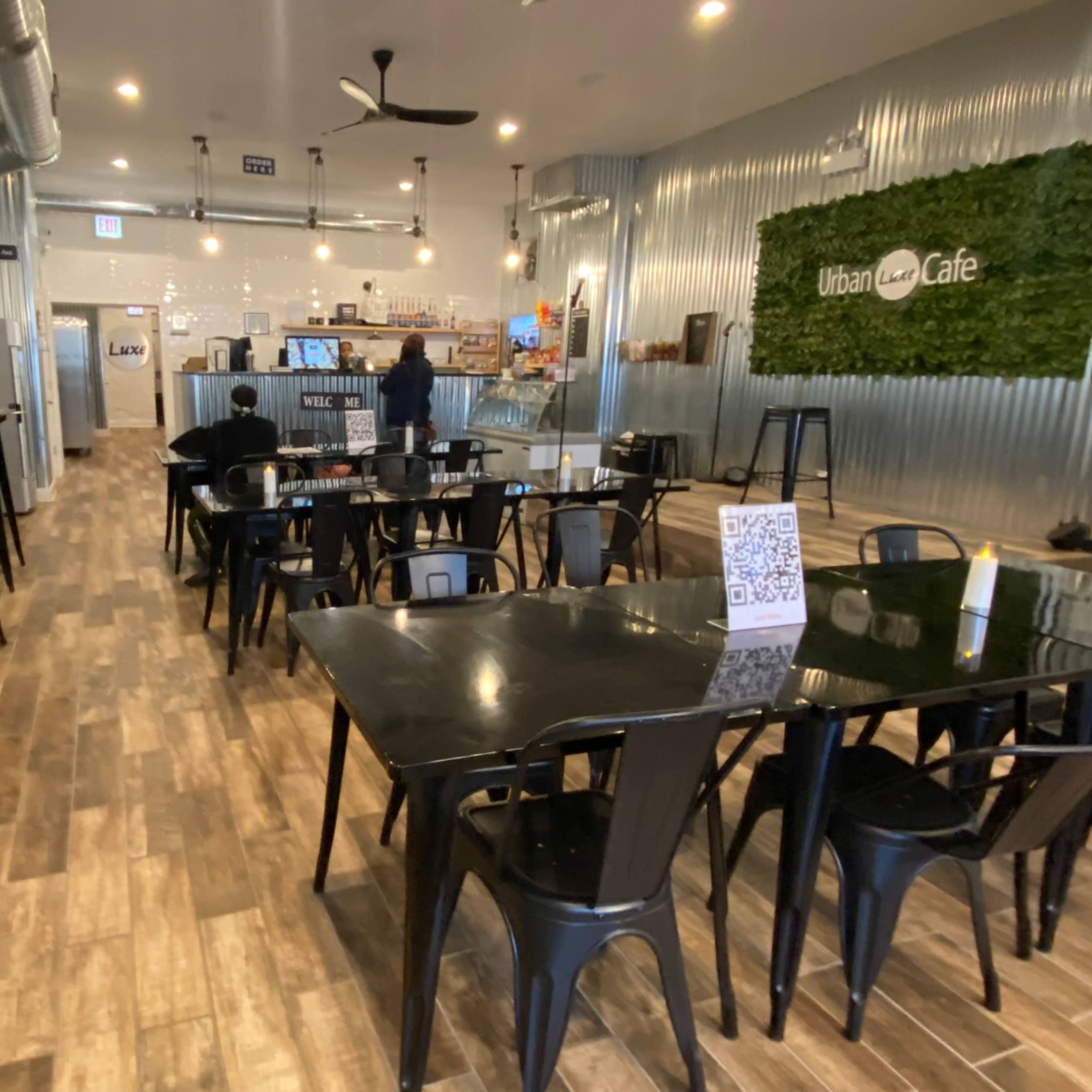 The interior of a café features black tables and chairs arranged neatly on a wooden floor, with a counter and kitchen area visible in the background.