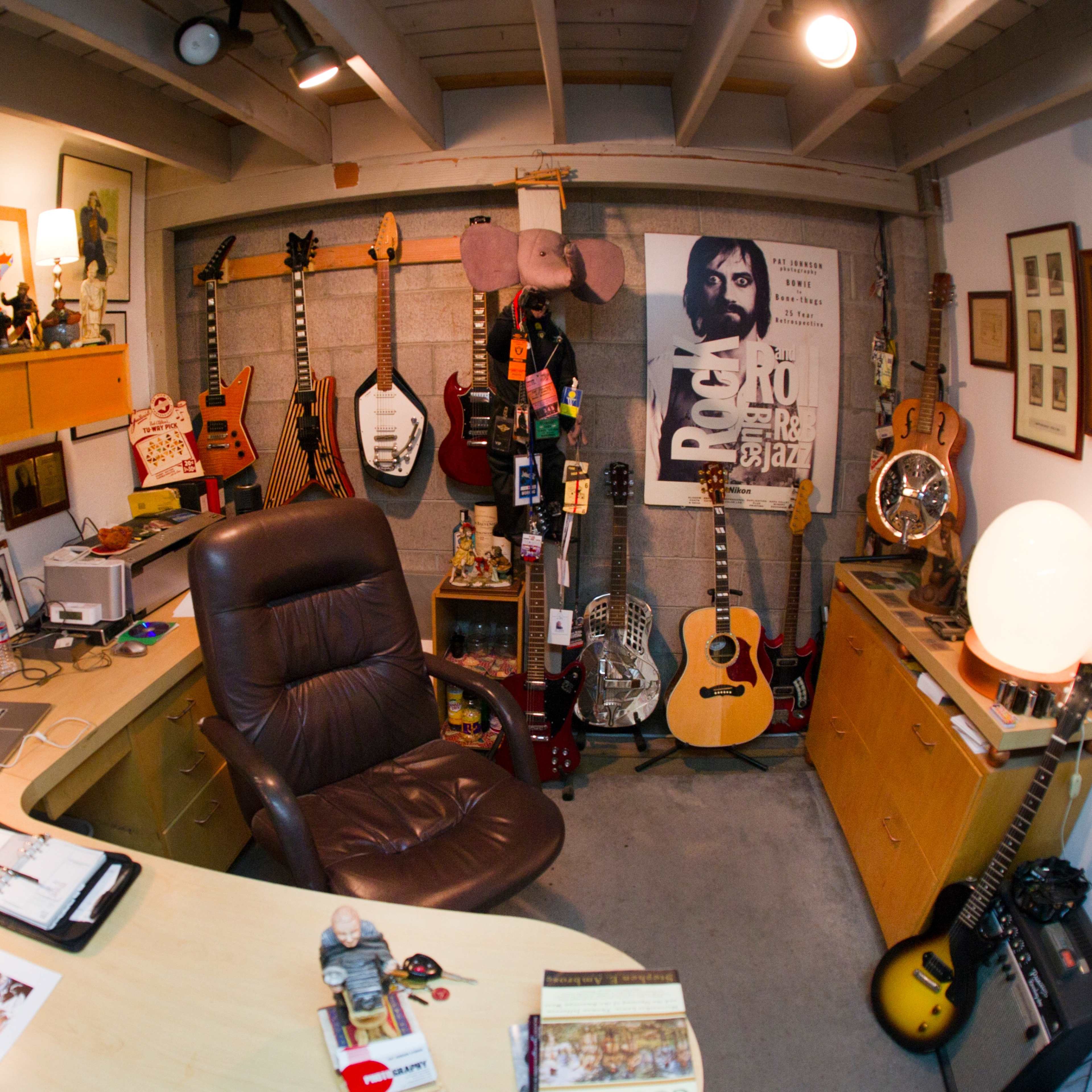 A cluttered home office filled with various guitars, memorabilia, and a desk with a computer chair.