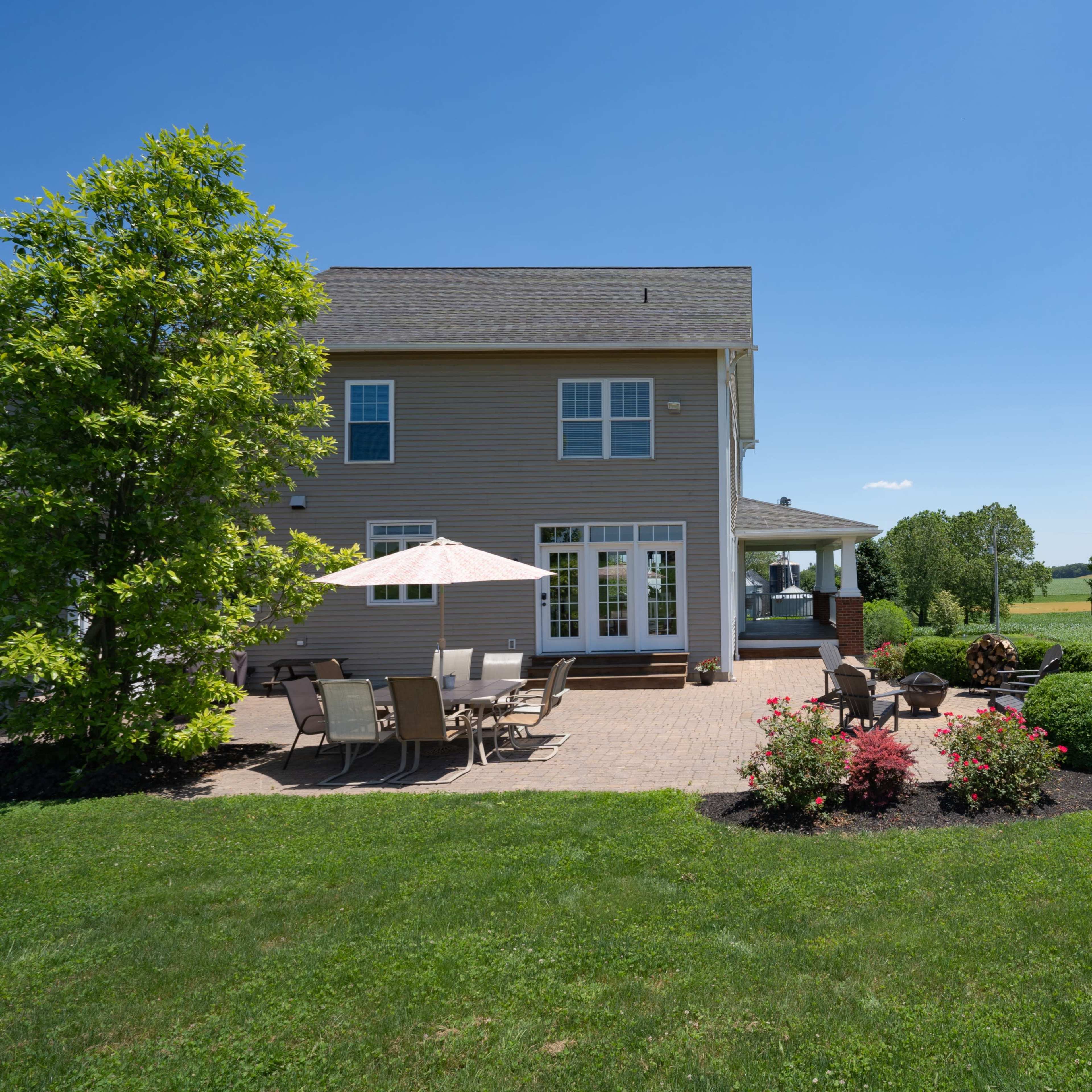 A two-story house features a patio area with outdoor seating and a view of a green field under a clear blue sky.
