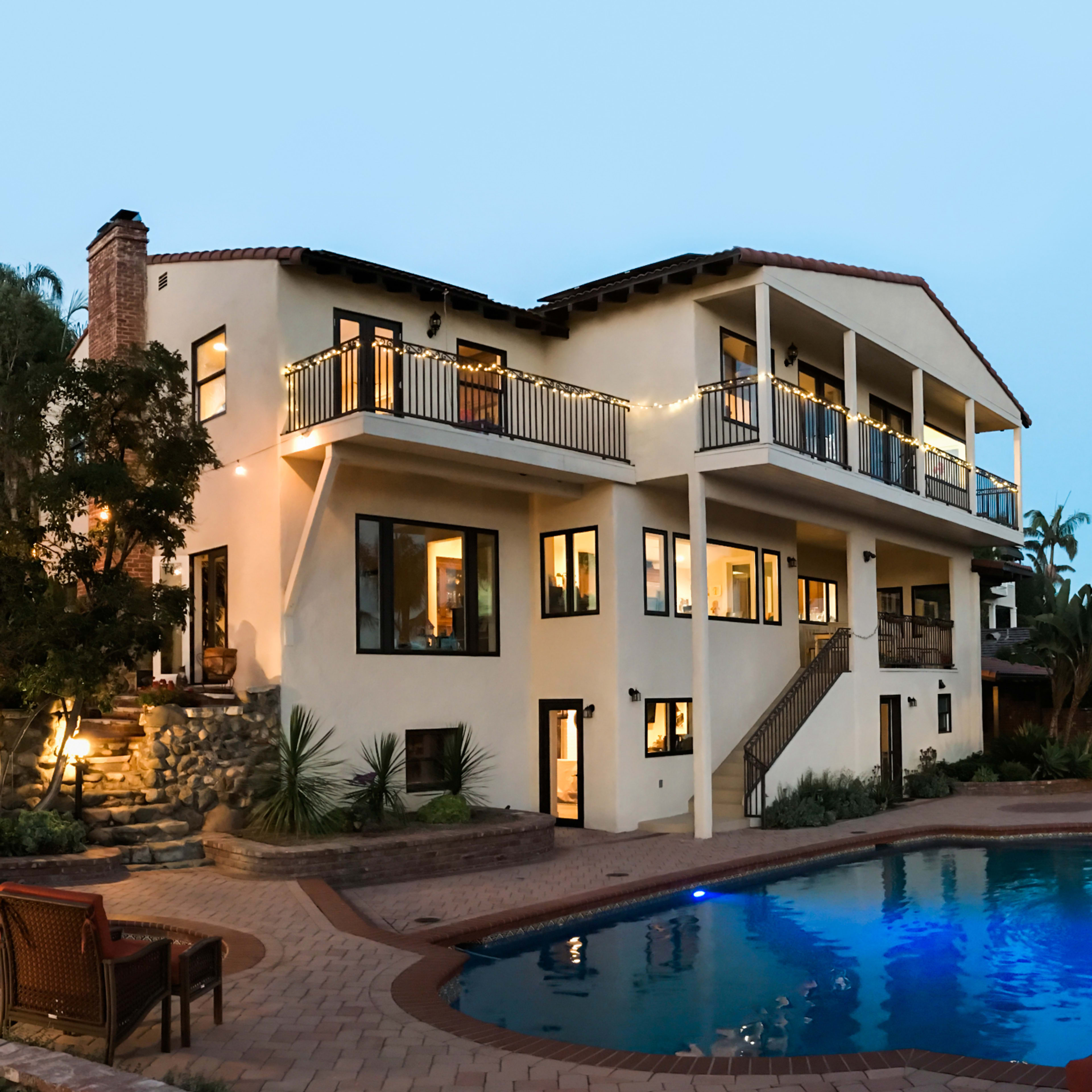 A two-story house with large windows and a balcony overlooking a blue swimming pool surrounded by palm trees and outdoor seating areas.
