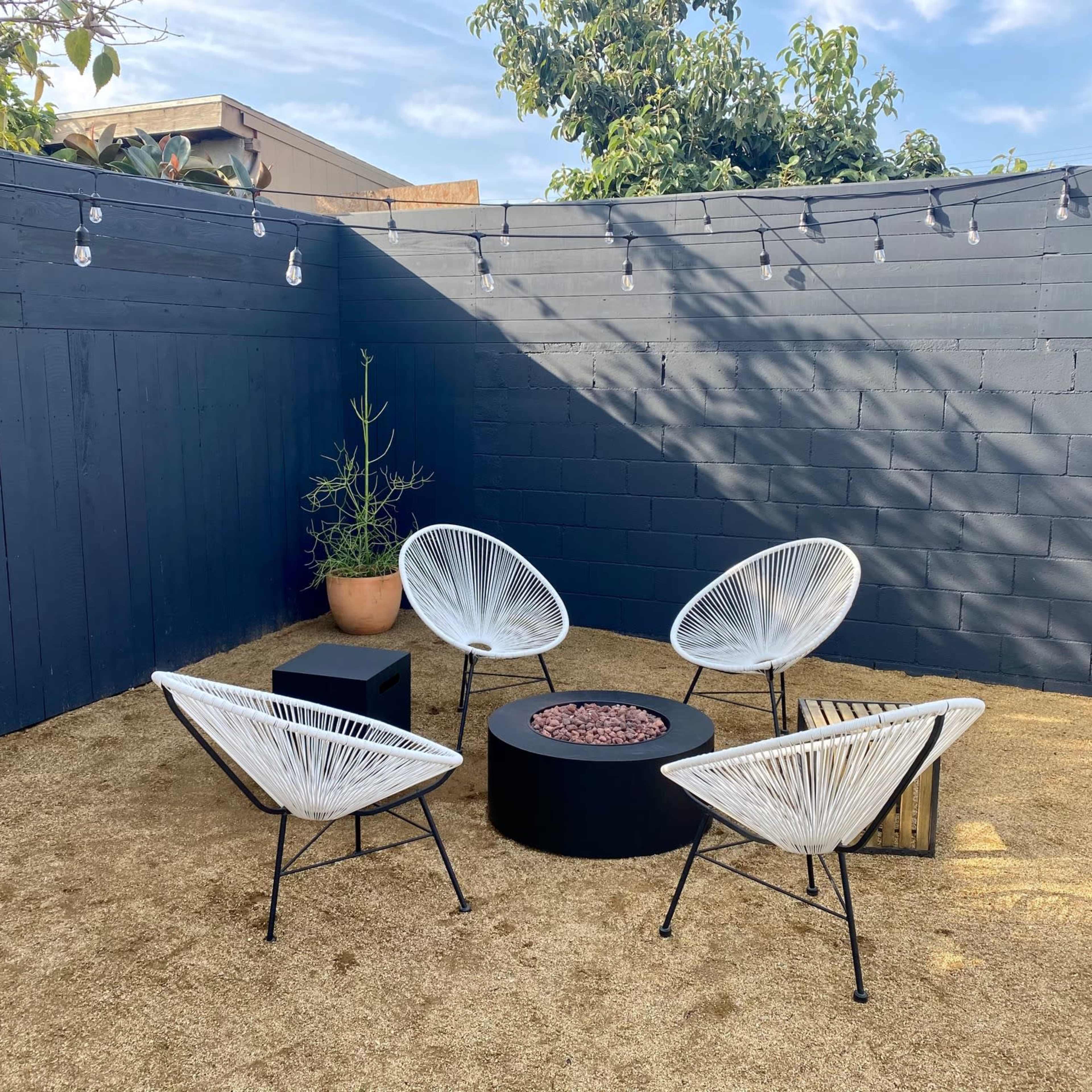 A small outdoor space with four white chairs arranged around a black fire pit on a sandy surface, enclosed by a blue wall and adorned with string lights overhead.