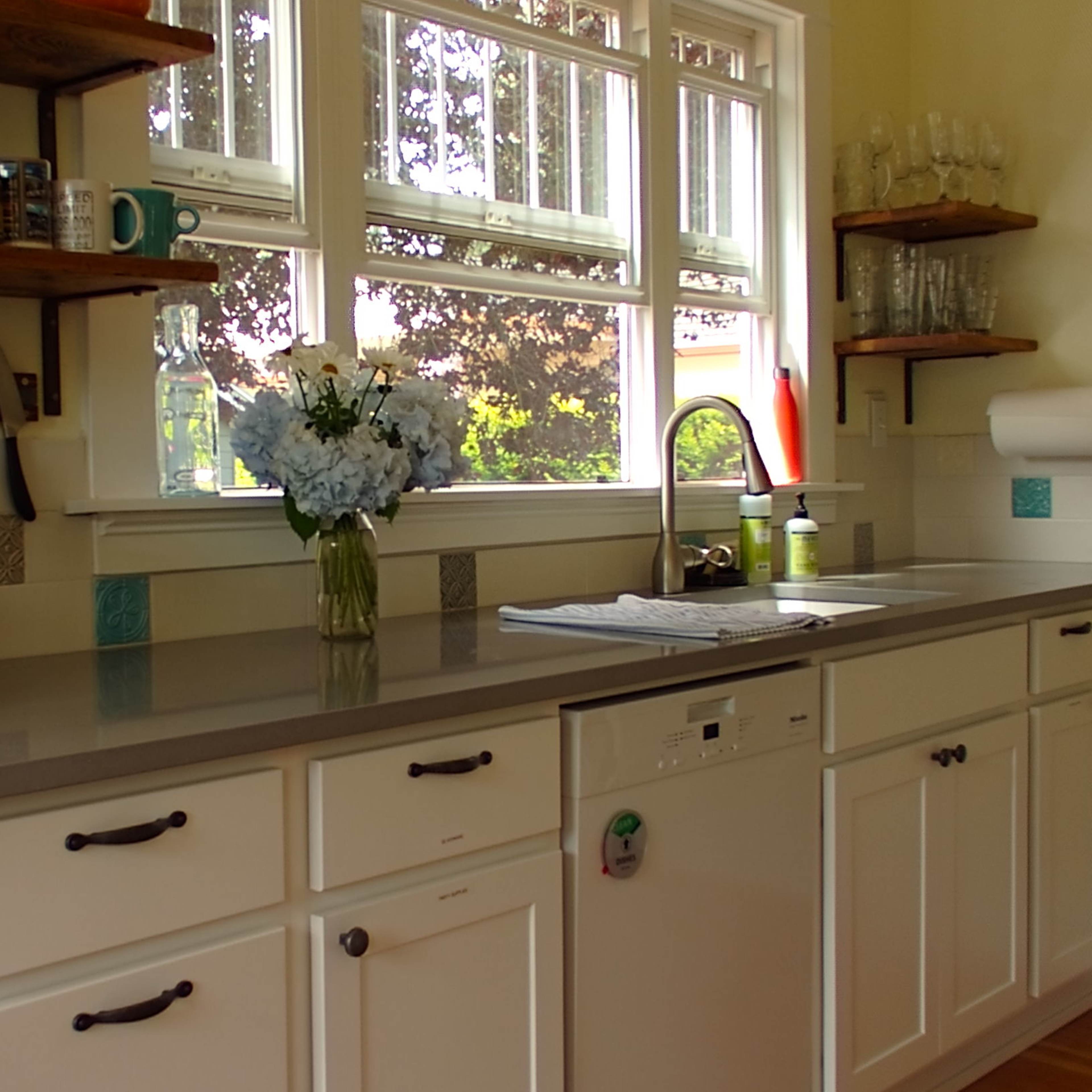 A kitchen with a countertop, a coffee maker, a vase of flowers, and shelves holding various kitchenware and glassware.