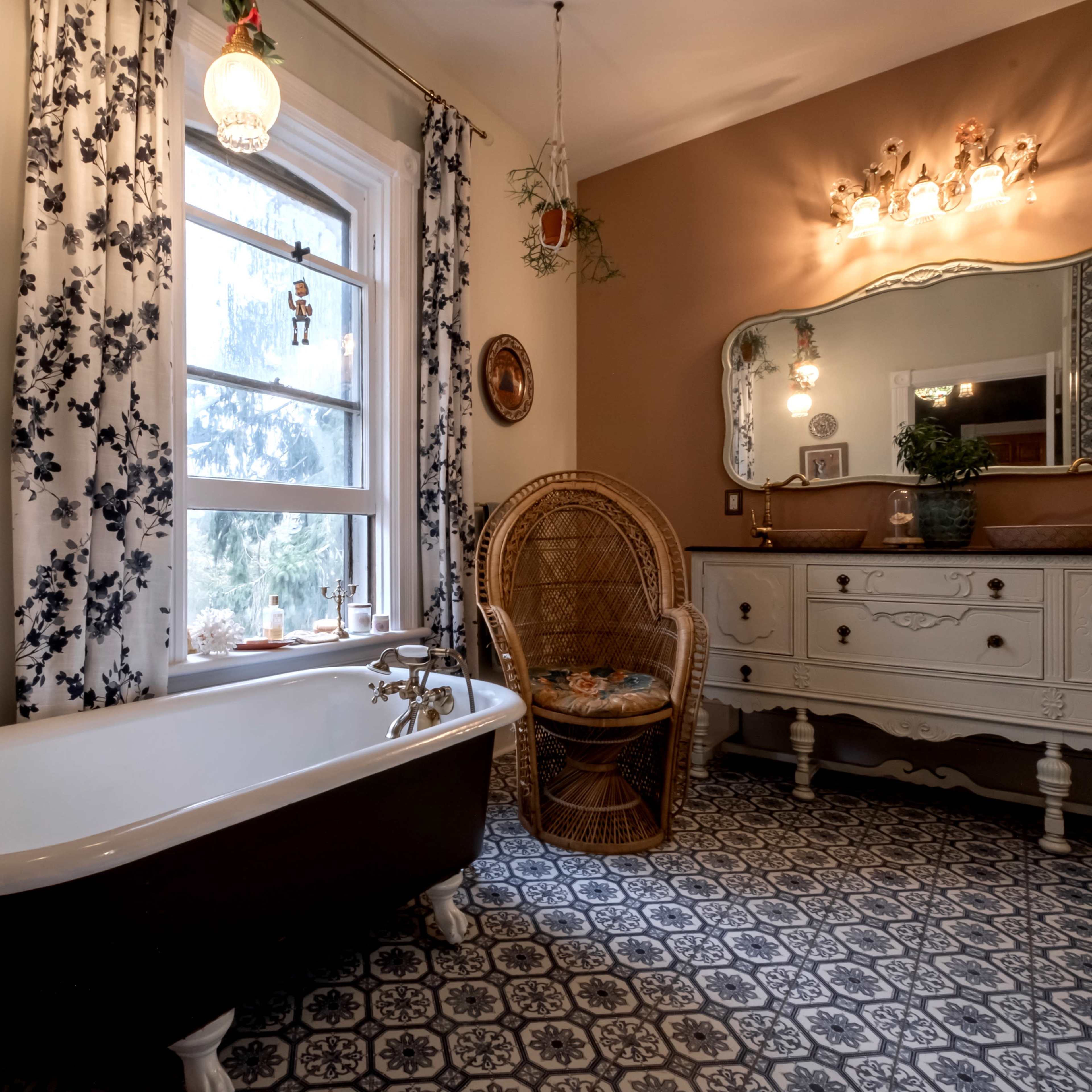 A bathroom featuring a freestanding clawfoot bathtub, a wicker chair, and a vintage-style dresser, with floral curtains framing a window.