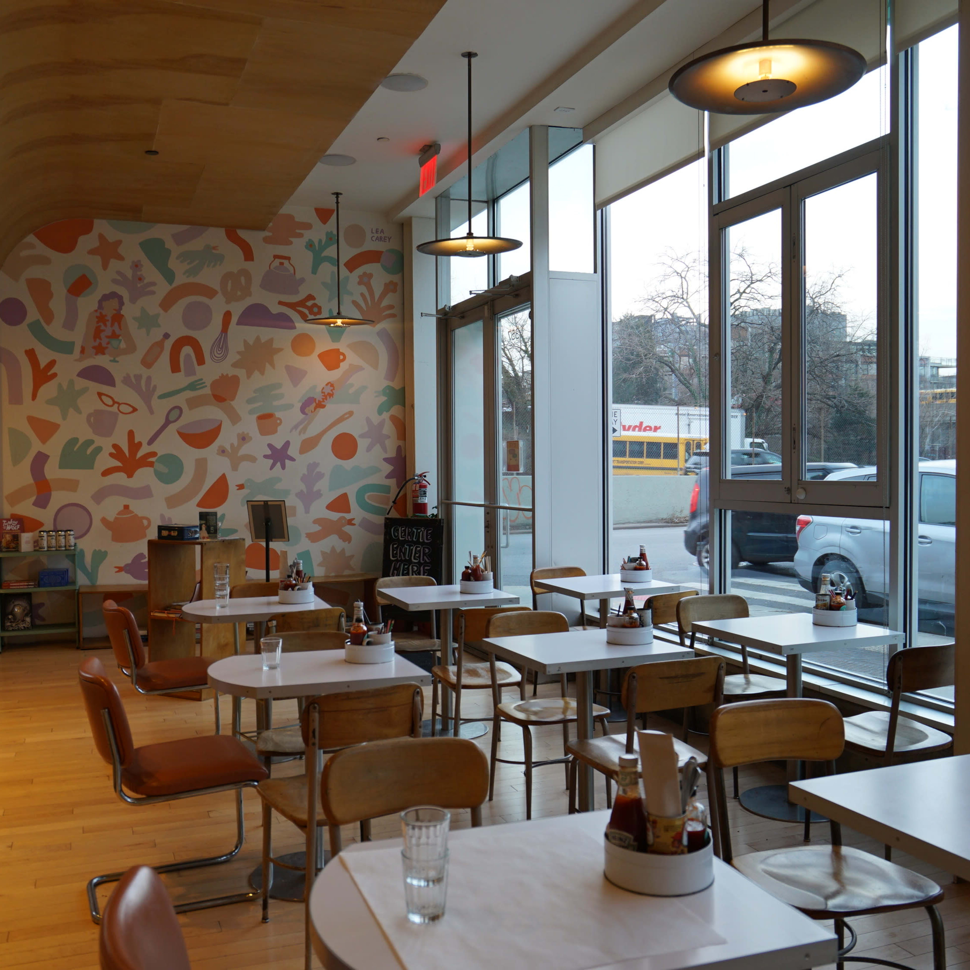 A modern café interior with wooden accents, colorful wall decor, and a row of tables arranged near large windows.