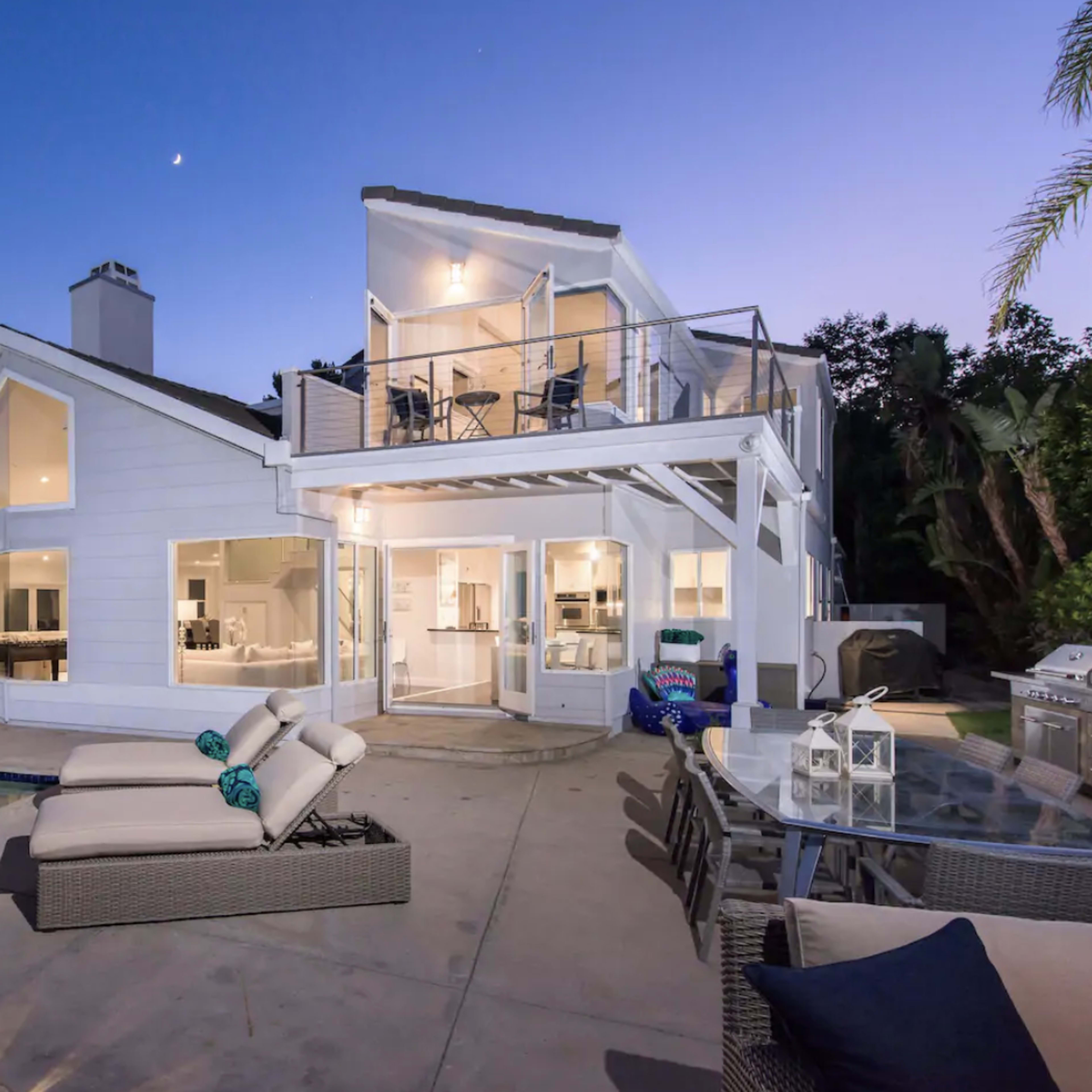 A modern white house with large windows and a balcony overlooks a swimming pool in a twilight setting.