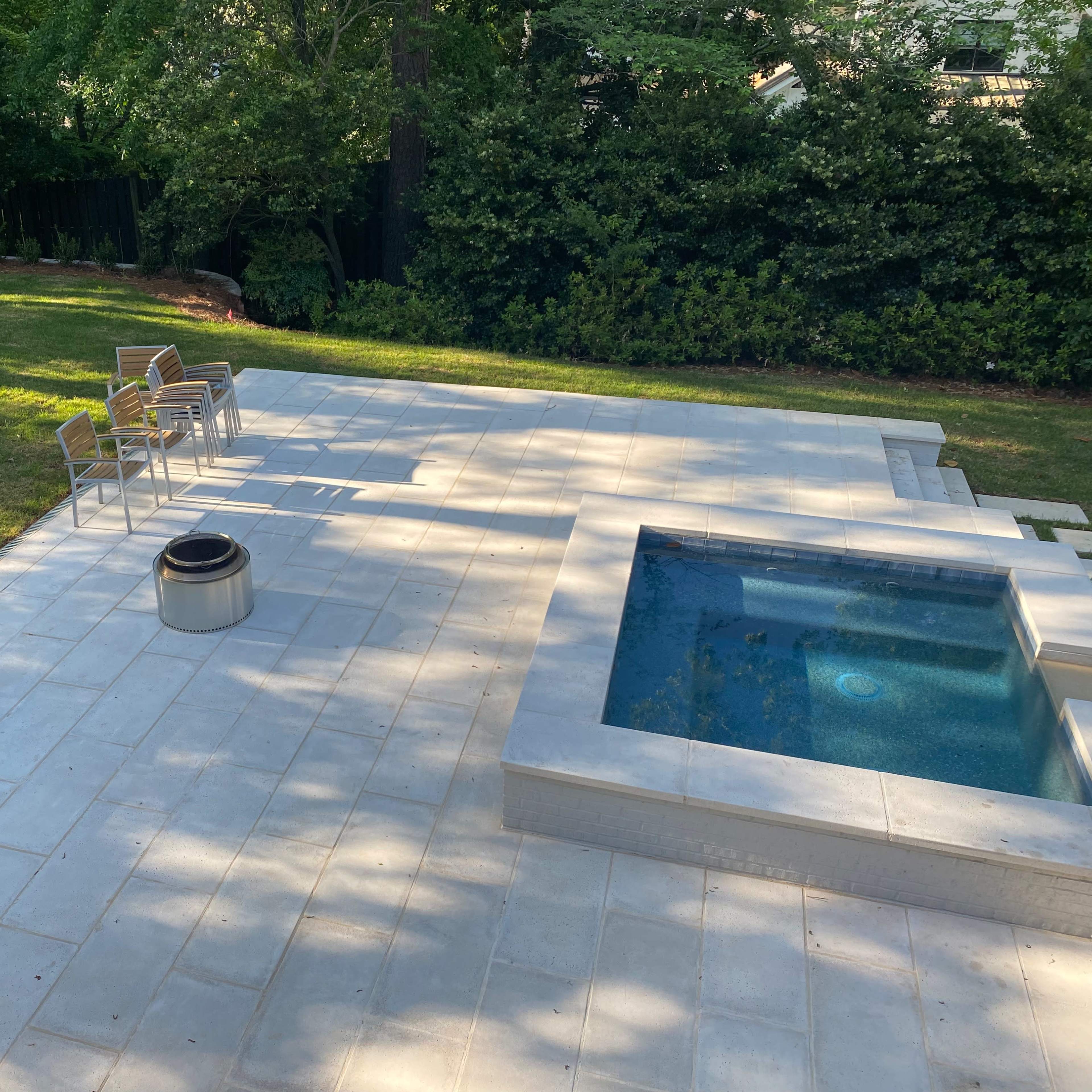 A spacious patio with a small outdoor spa, a fire pit, and several wooden chairs arranged nearby on a lawn.