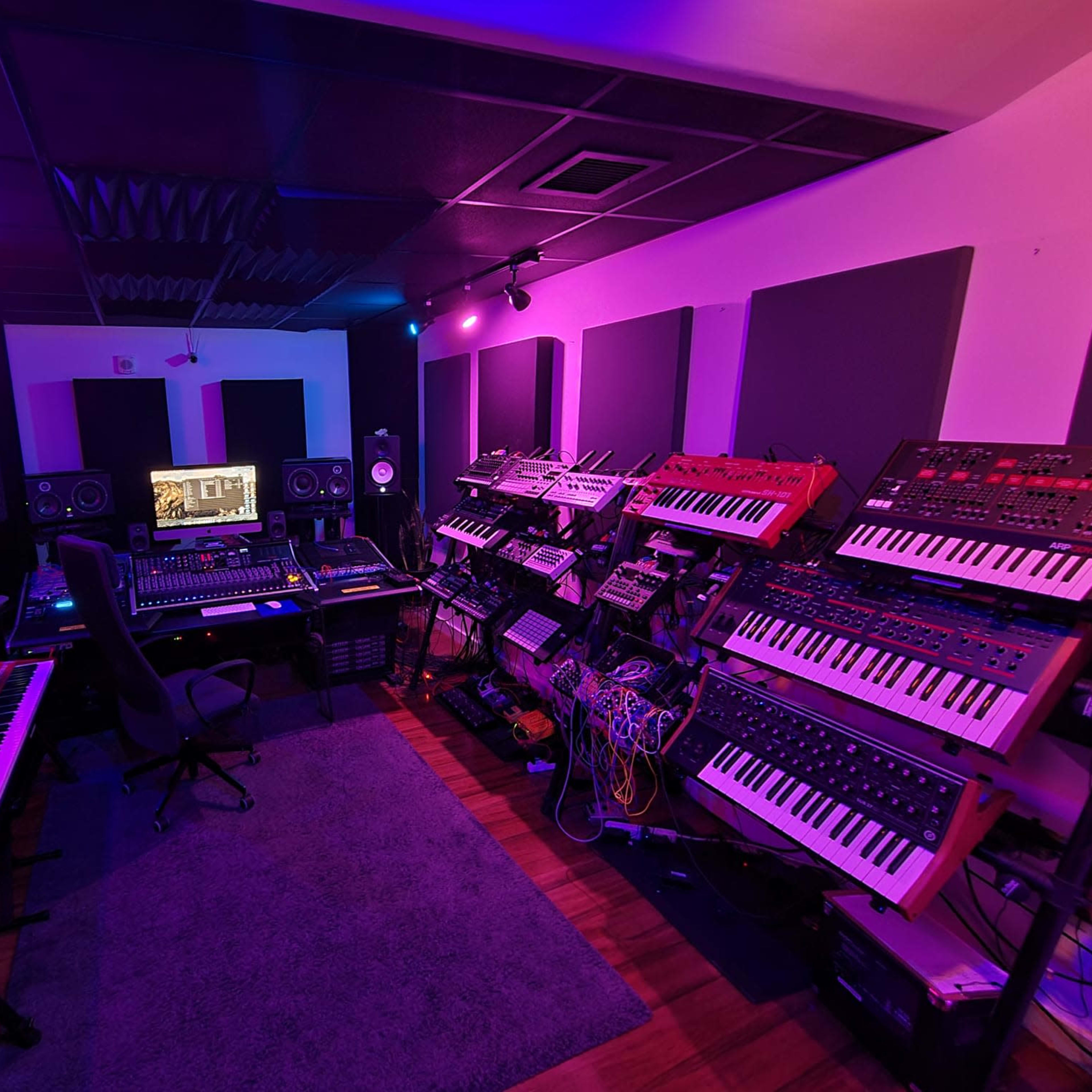A music studio equipped with various synthesizers, a mixing console, and workstations, surrounded by soundproof panels.
