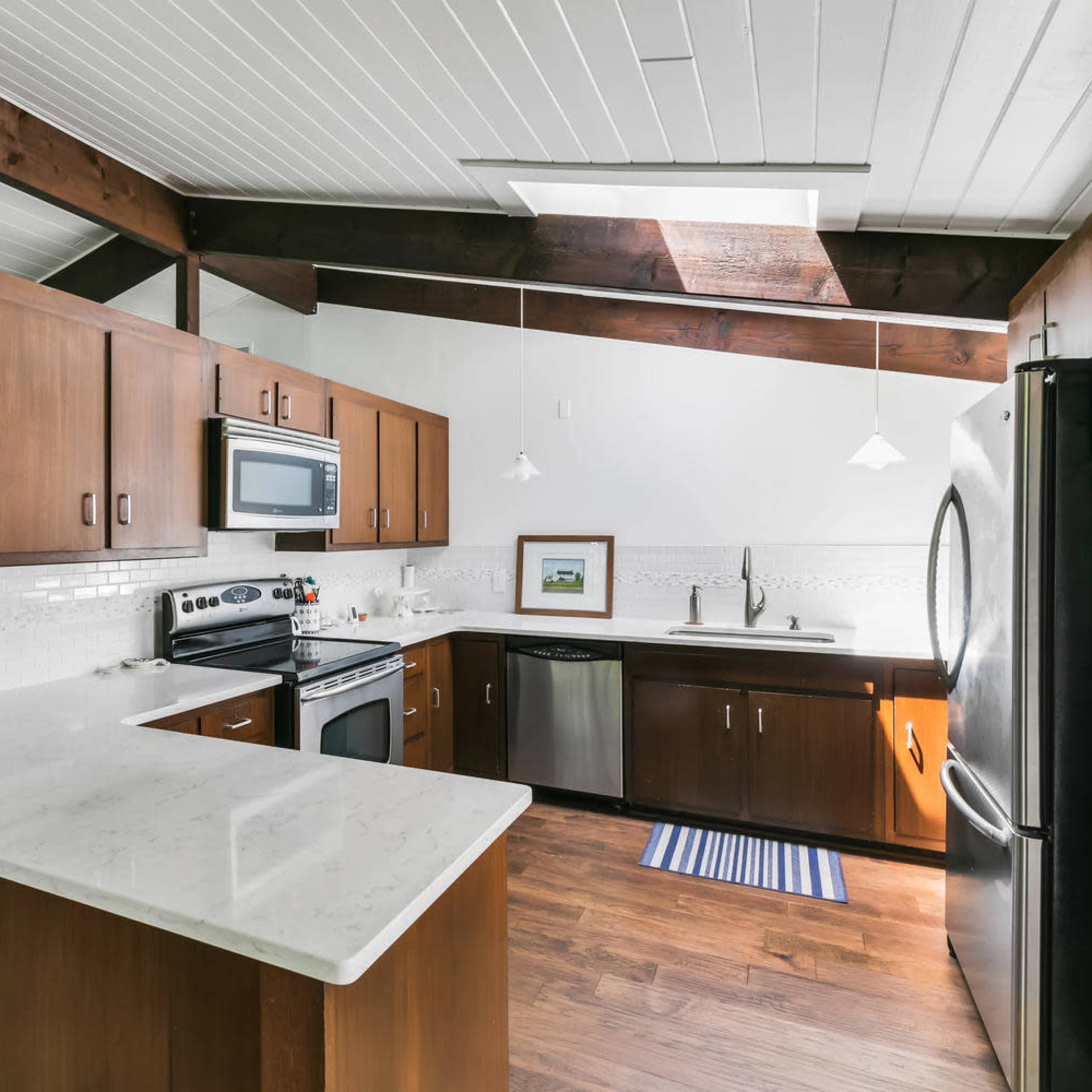 A modern kitchen features dark wood cabinetry, stainless steel appliances, and a white marble countertop beneath vaulted beams.