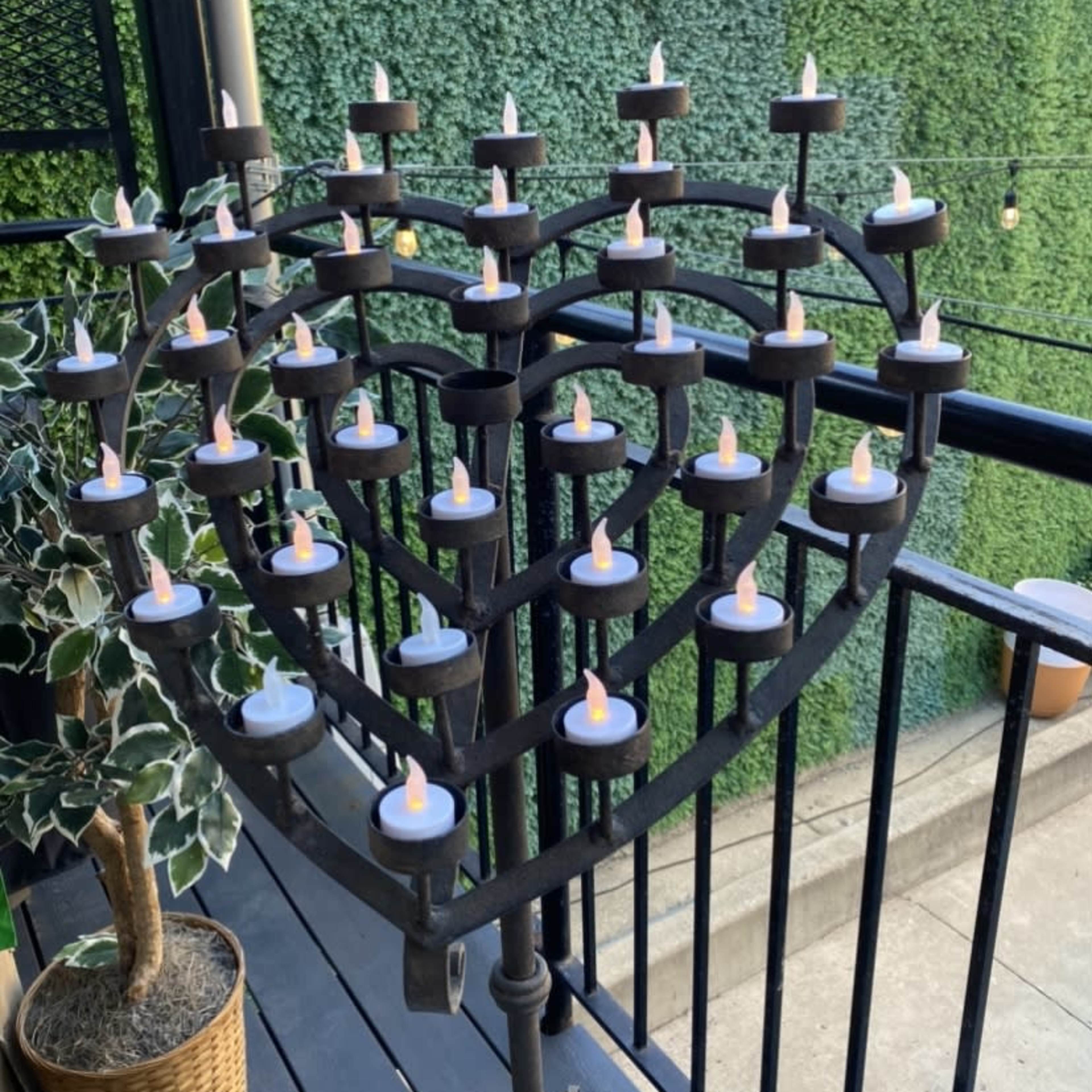 A heart-shaped metal stand holds multiple candles on a balcony surrounded by greenery.
