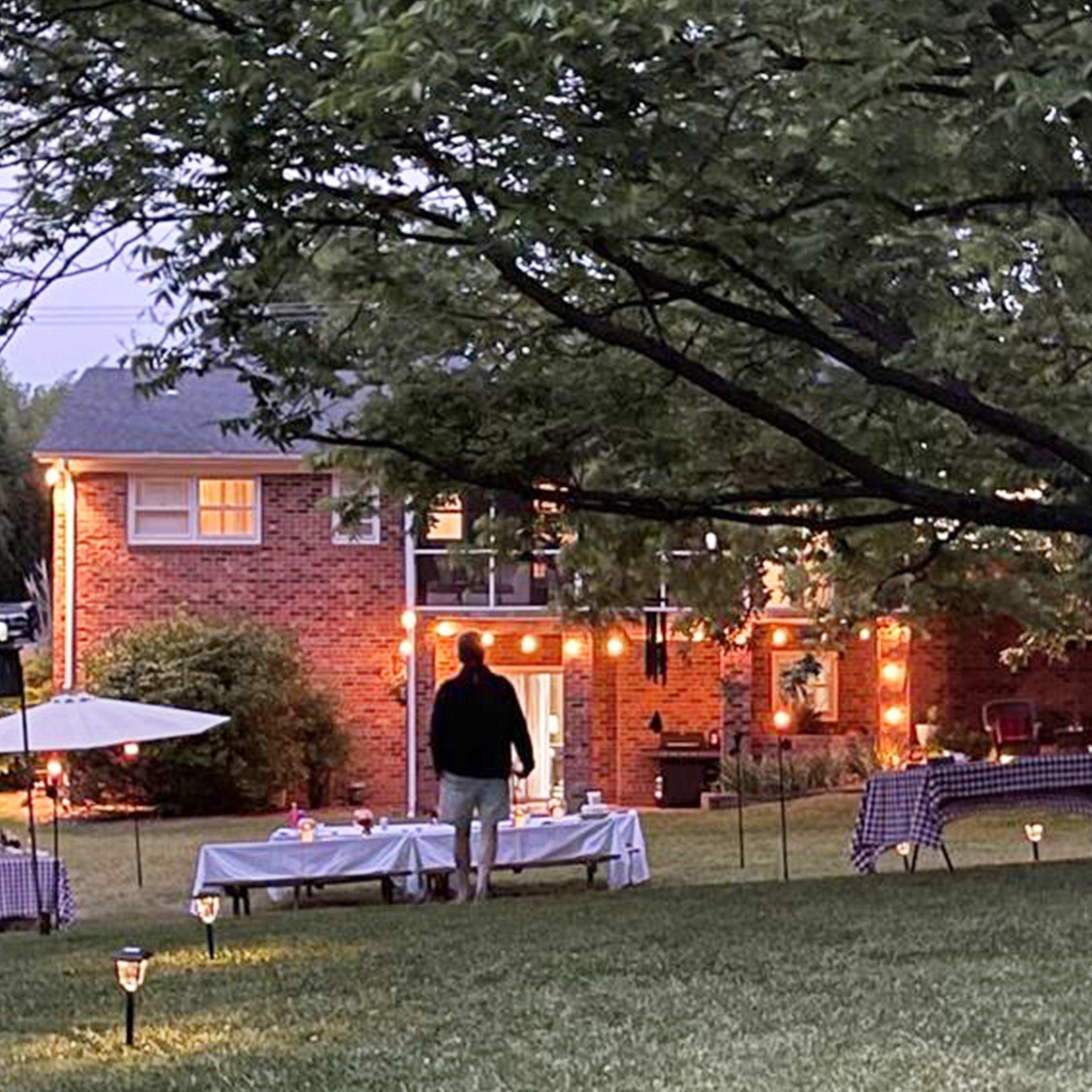 A man stands near outdoor tables illuminated by string lights in a backyard with a brick house in the background.