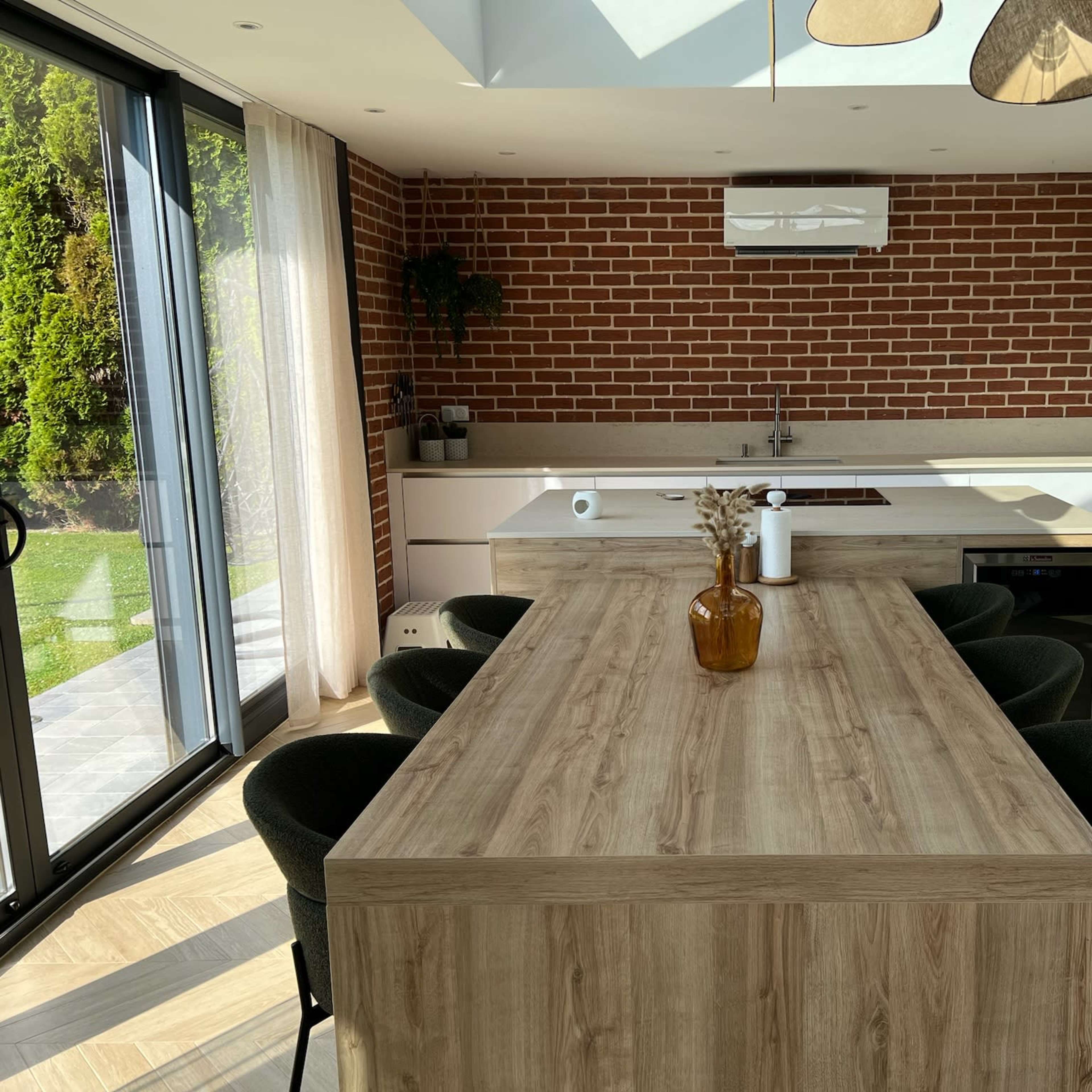 A modern kitchen with a wooden dining table, brick walls, and large glass doors leading to an outdoor area.