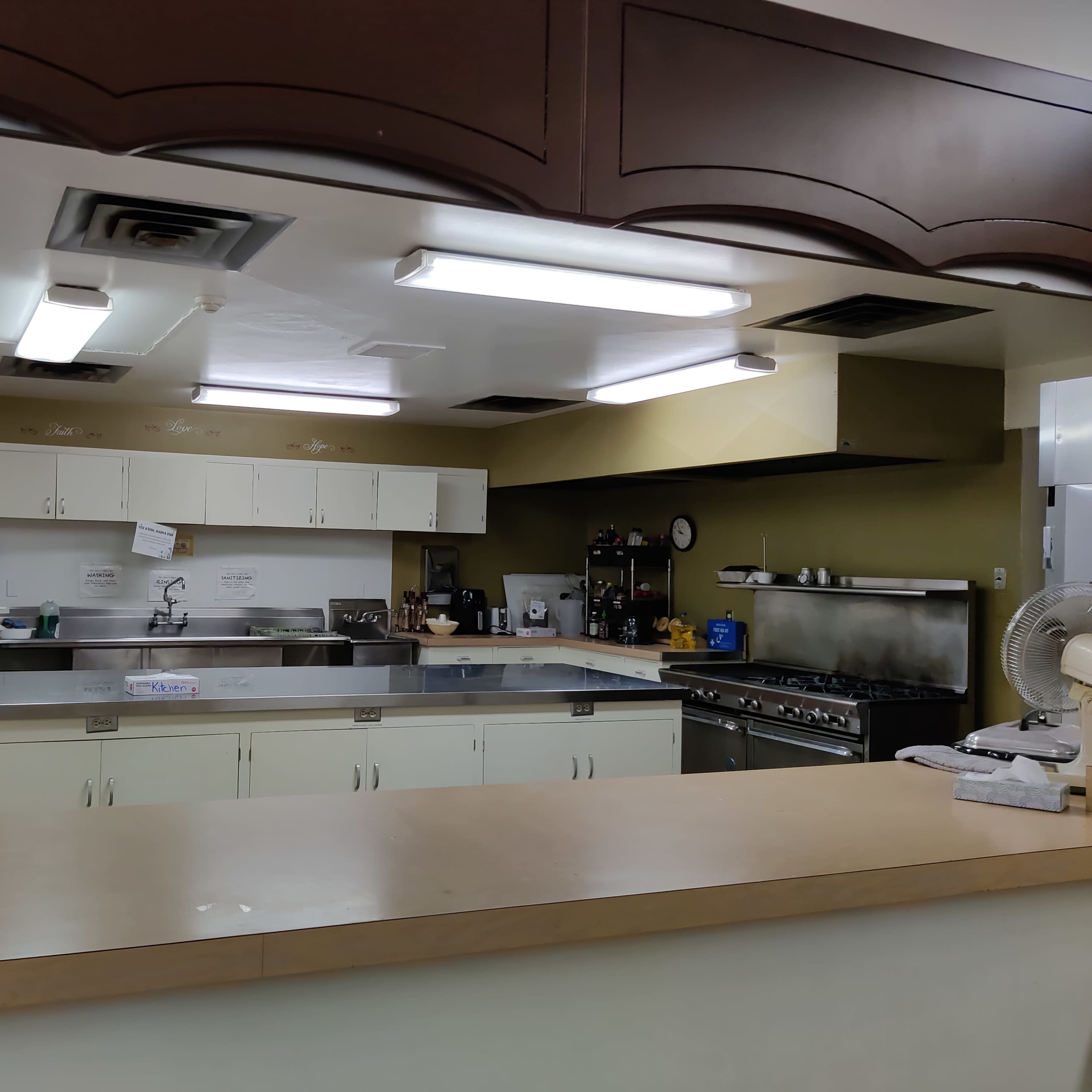 A kitchen with a counter in the foreground, featuring multiple appliances and workspaces under fluorescent lighting.