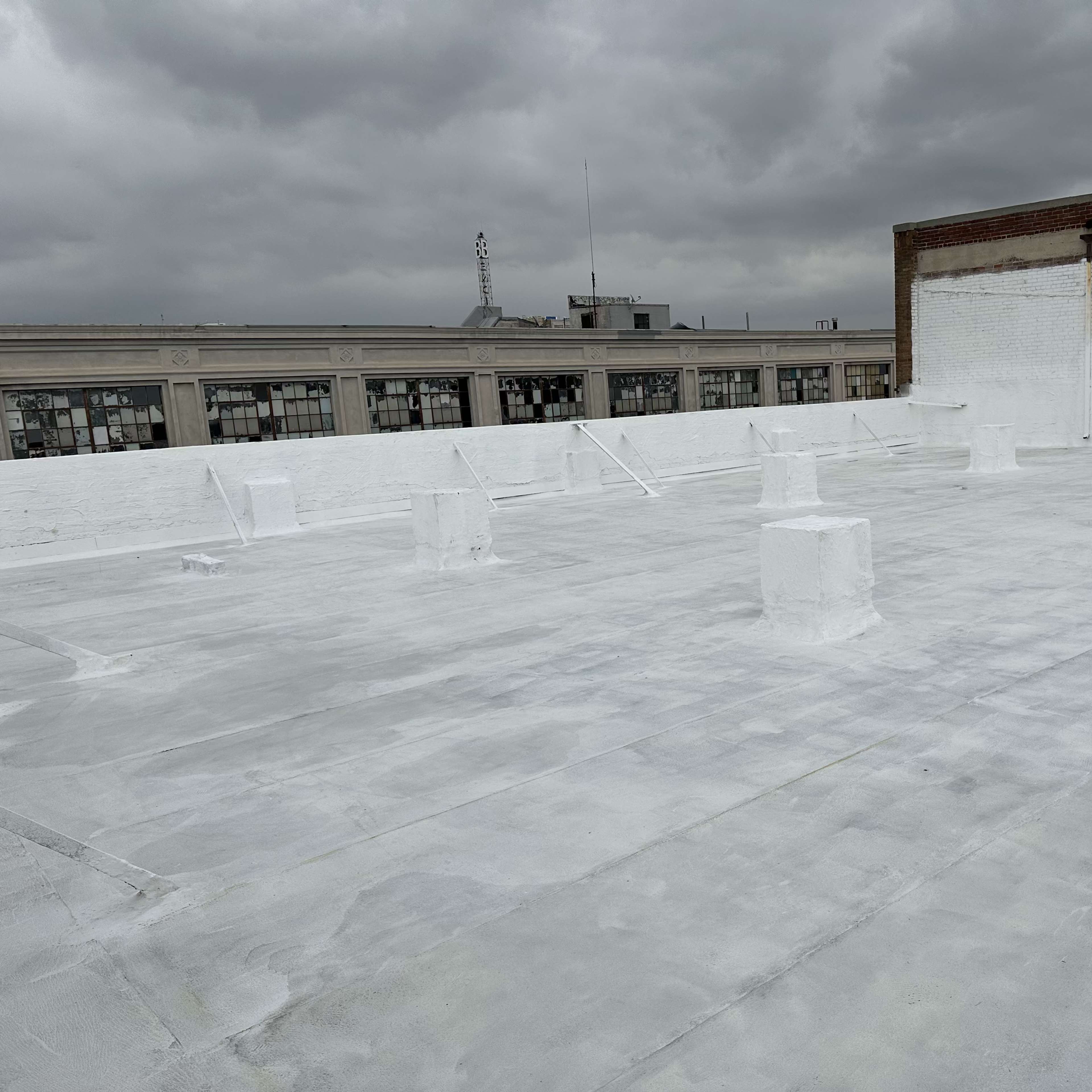 A flat rooftop covered in a white coating, featuring several cylindrical structures and a large building in the background under a cloudy sky.