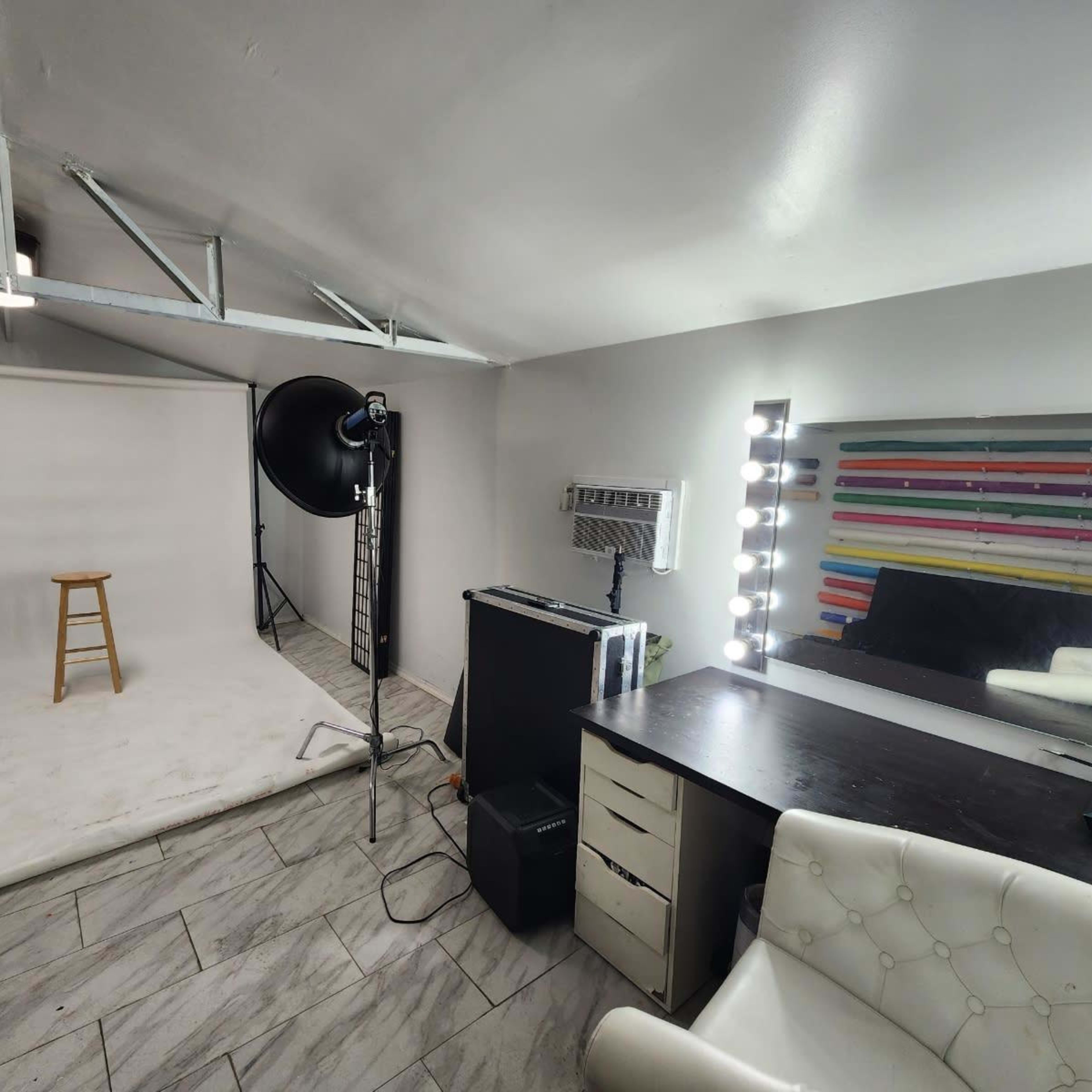 A photography studio with a white backdrop, a stool, lighting equipment, and a vanity mirror with lights.