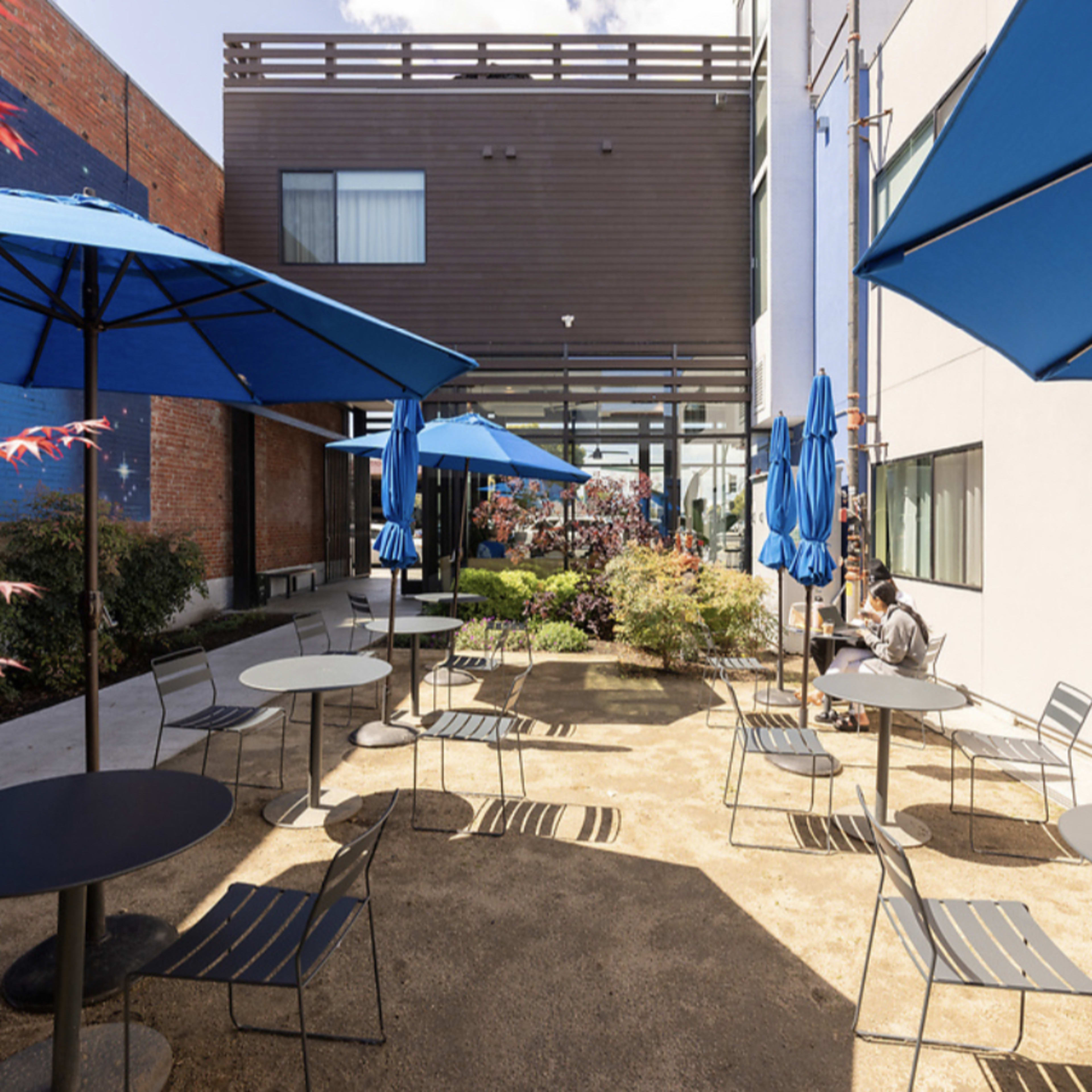 A courtyard features several tables and blue umbrellas surrounded by greenery and modern buildings.