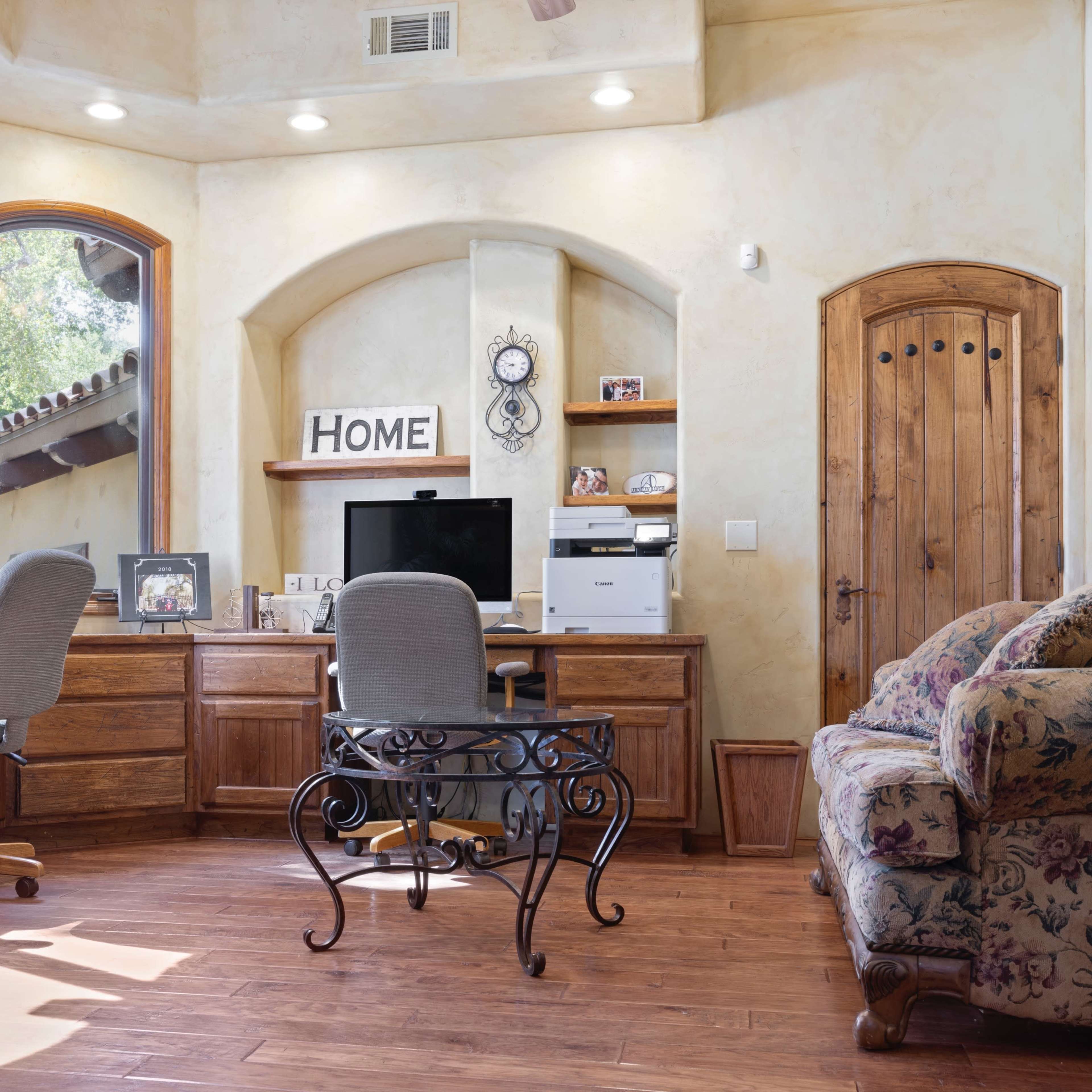 A home office features two chairs at a wooden desk with a computer, printer, and decorative elements in a well-lit room with large windows.