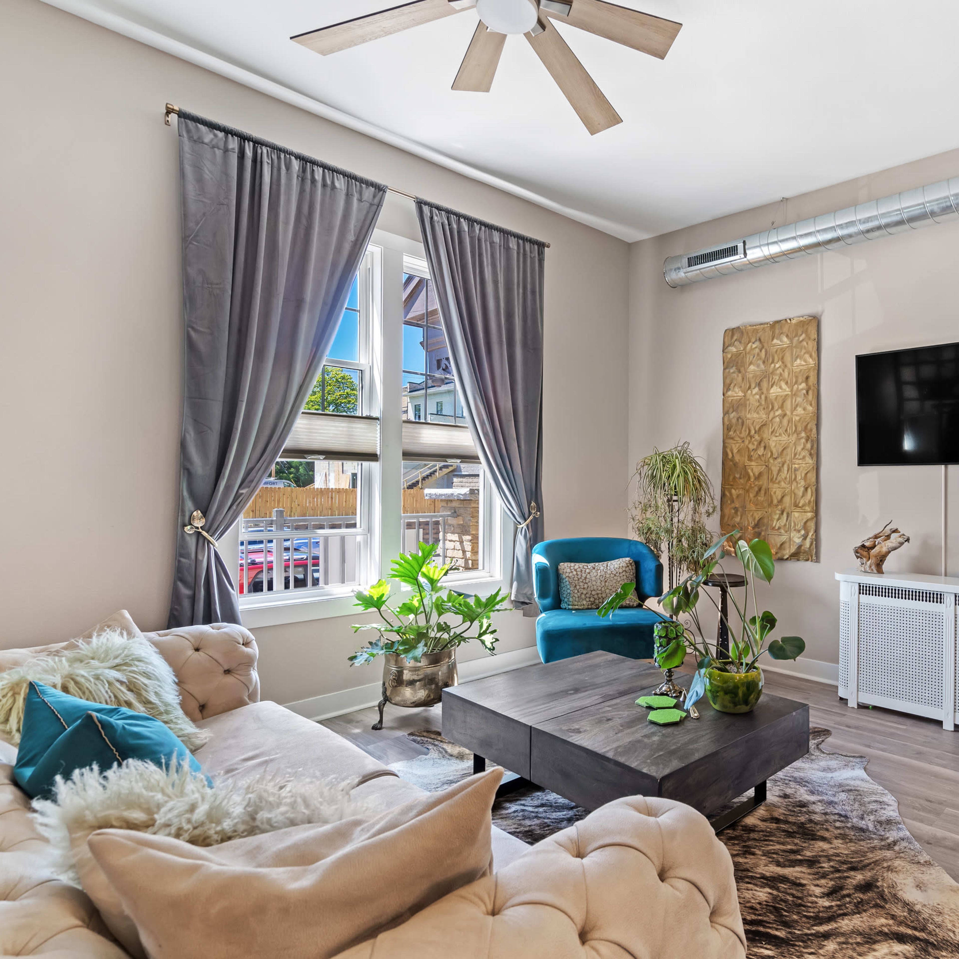 A cozy living room with a tufted sofa, a wooden coffee table, plants, and a mounted TV against a neutral backdrop.