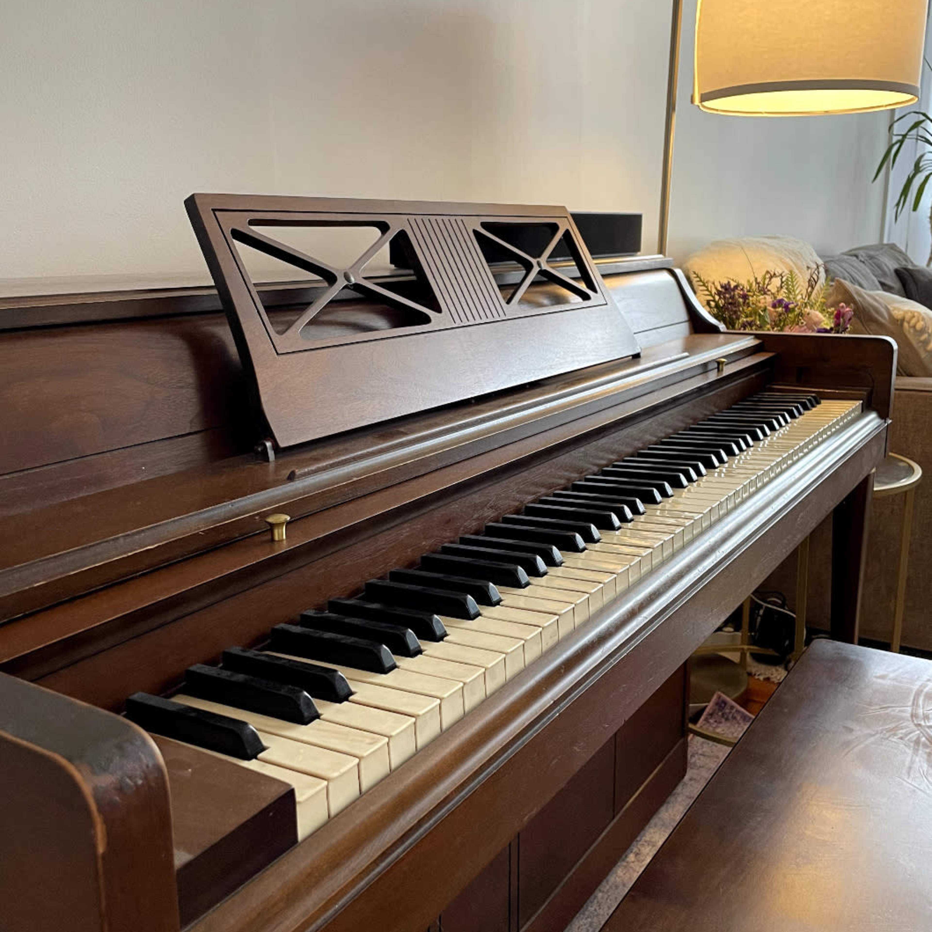 A wooden piano with a light-colored finish is positioned beside a lamp and a couch in a well-lit room.