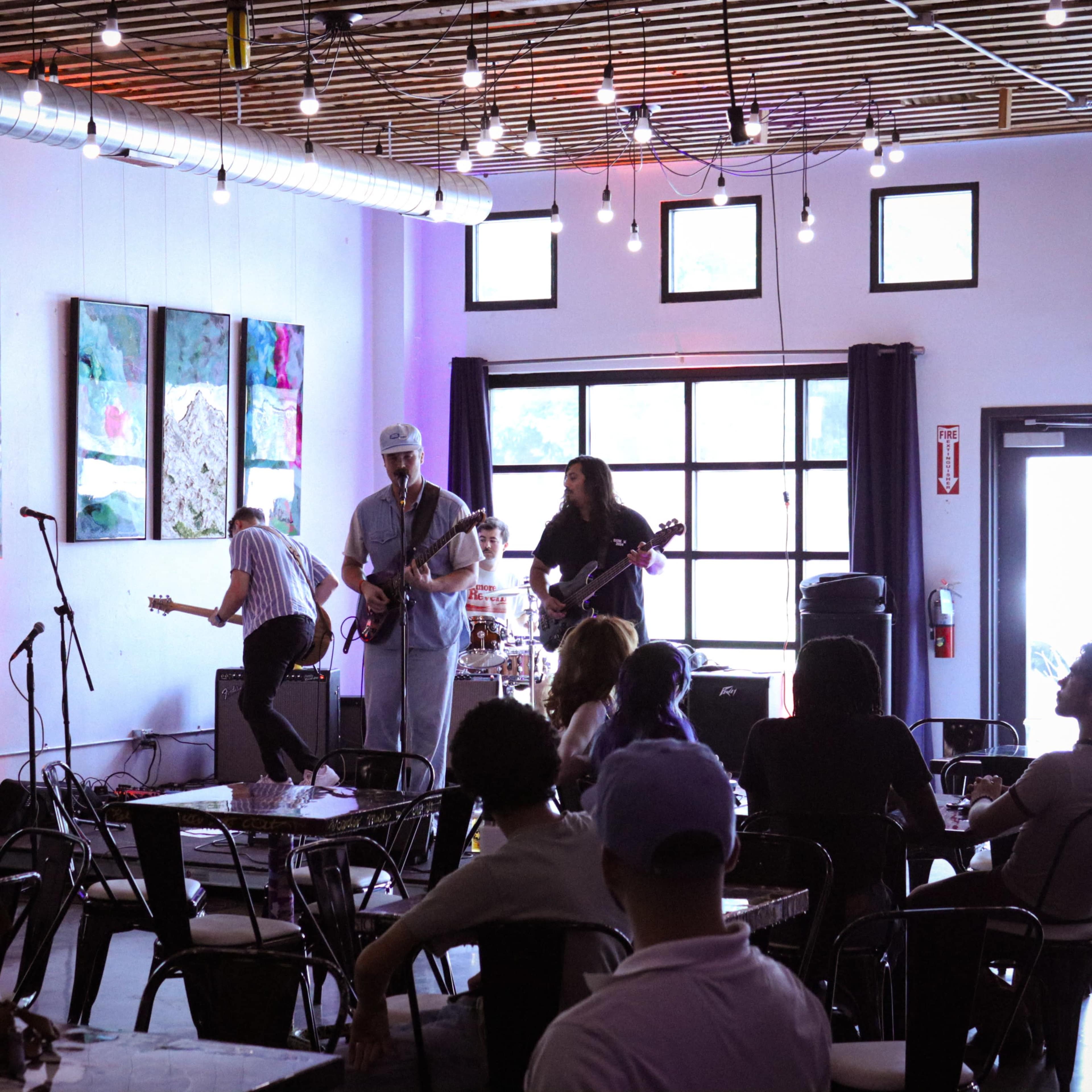 A live band performs onstage inside a modern café with a small audience seated at tables.