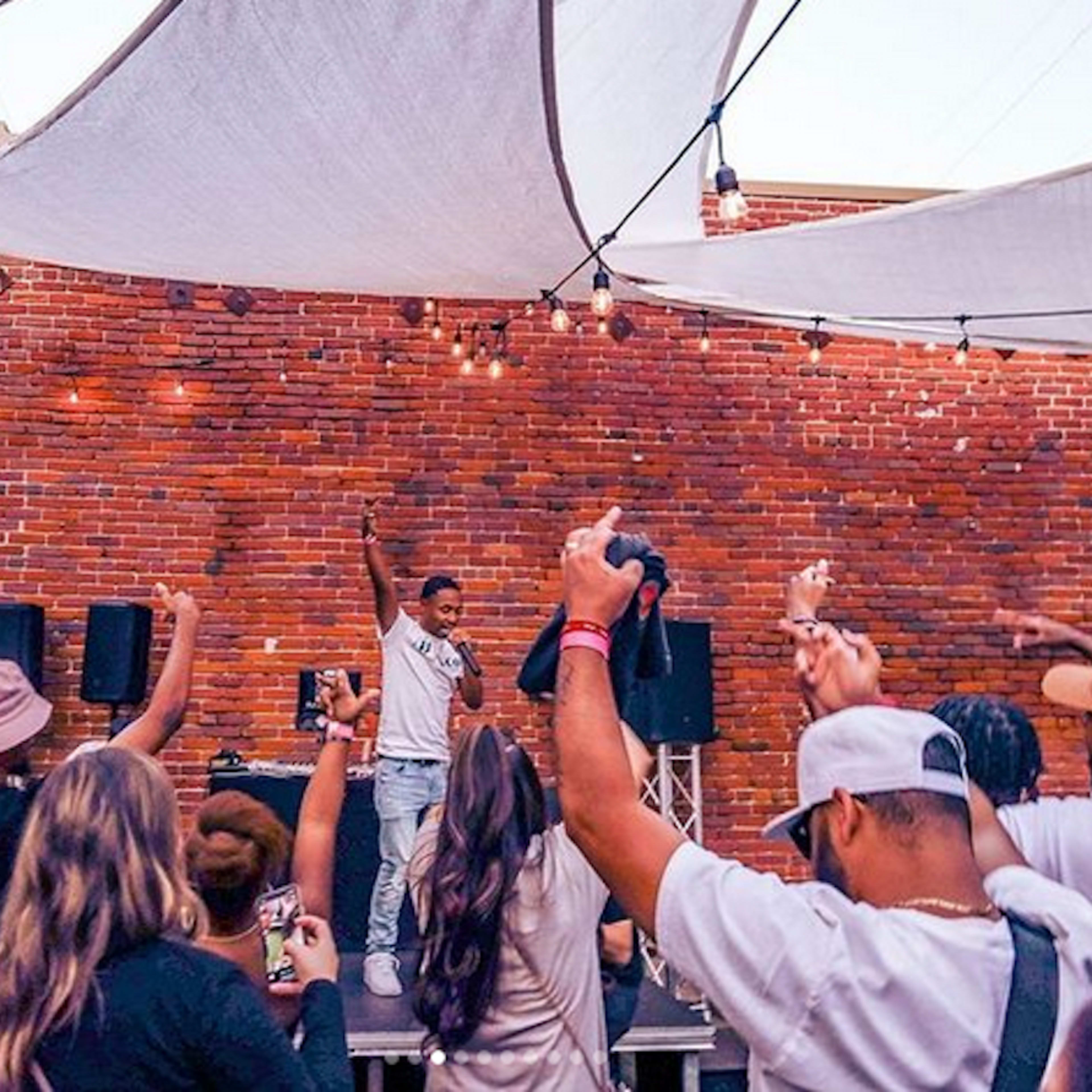 A performer stands on a stage, raising one hand while the audience, with their hands in the air, captures the moment on their phones under string lights and fabric canopies.