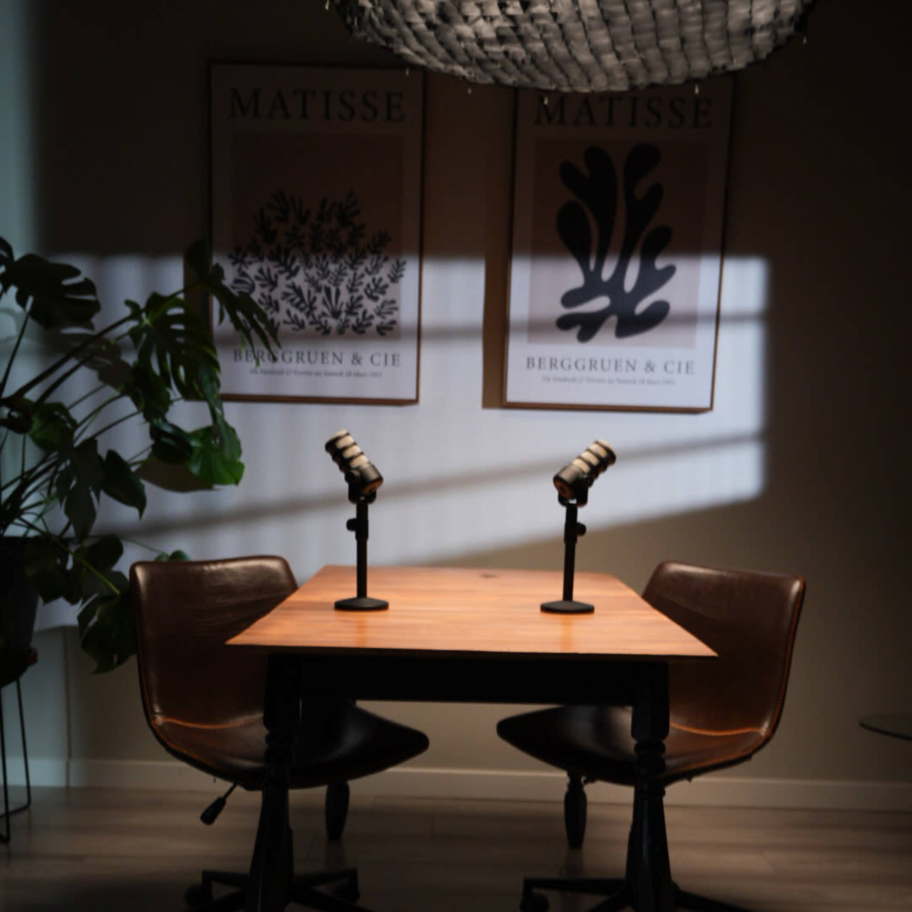 A wooden table with two chairs and microphones is set in a well-lit room, featuring two framed art pieces on the wall.