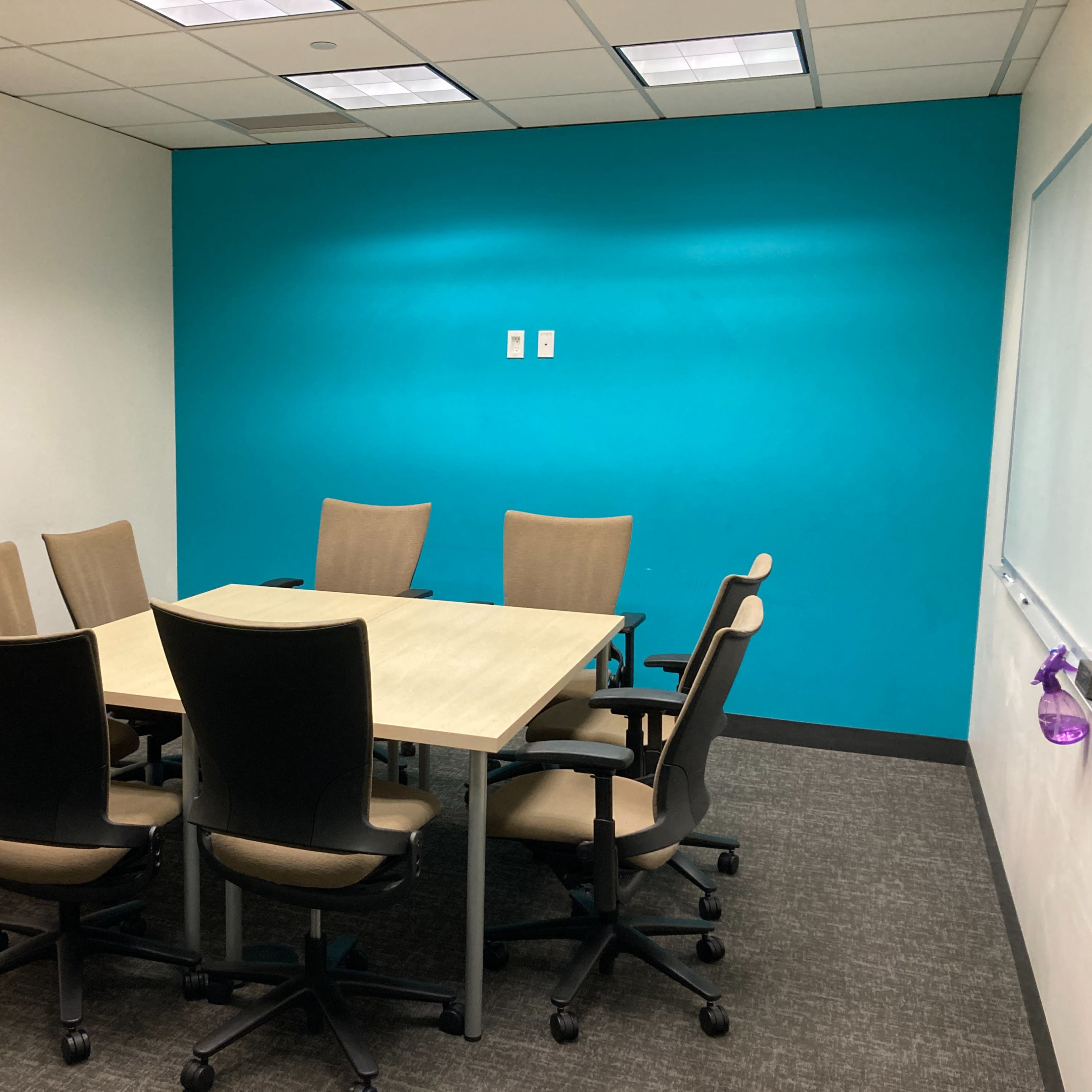 A conference room features a rectangular table surrounded by six chairs, with a bright turquoise wall and a whiteboard.