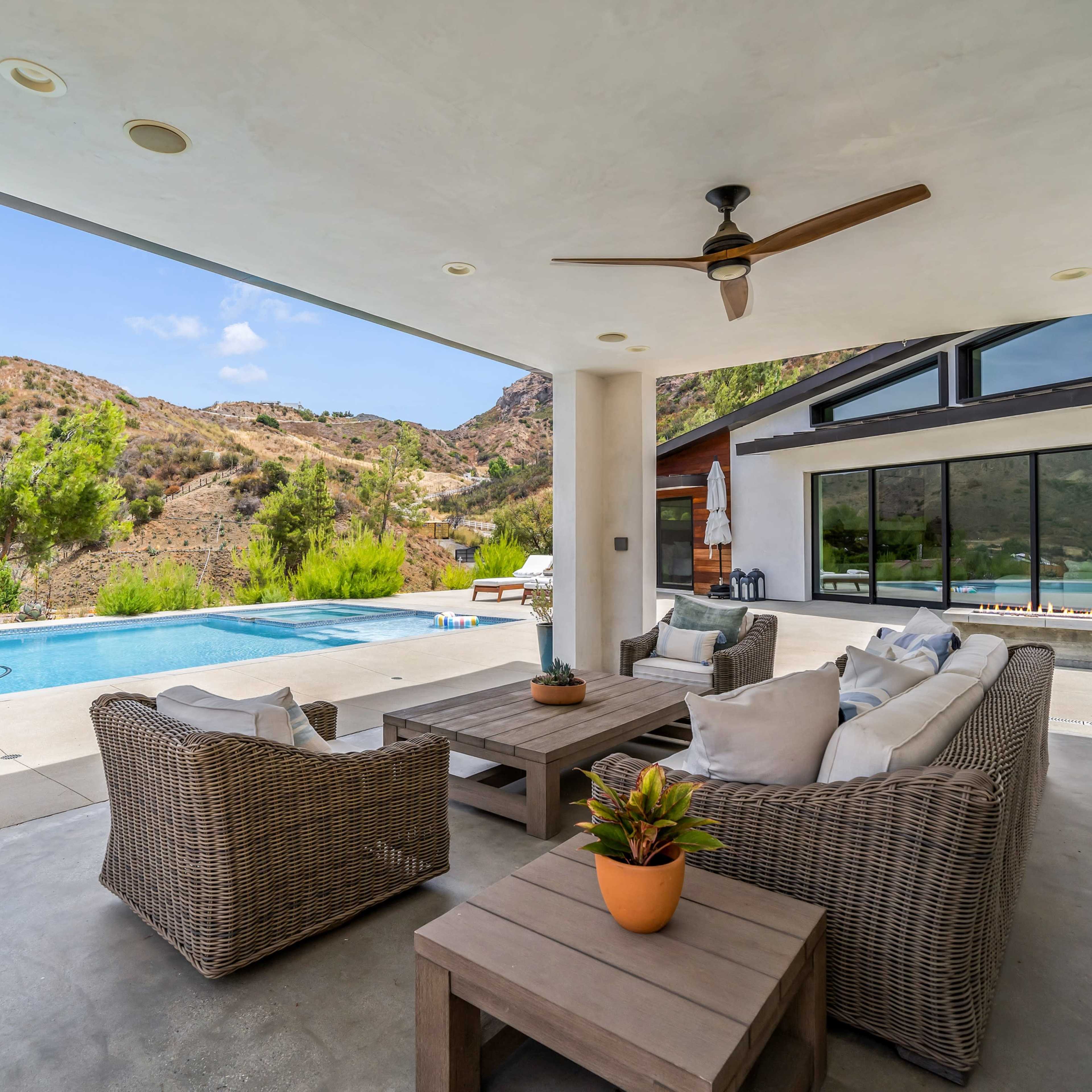 A covered patio features comfortable seating and a coffee table, overlooking a swimming pool and hills in the background.