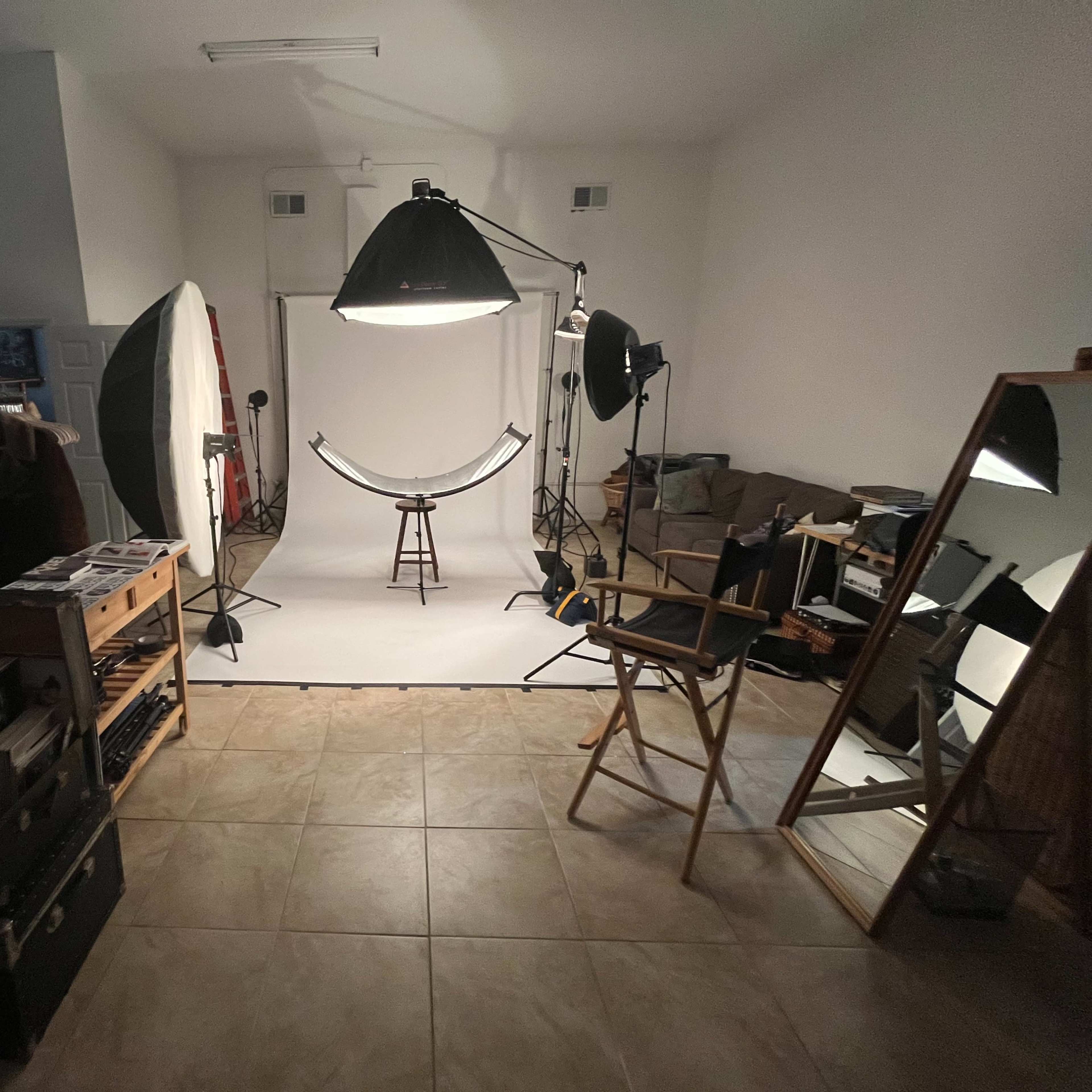 A photography studio featuring a white backdrop, various lighting equipment, a stool in the center, and a director's chair beside a large mirror.