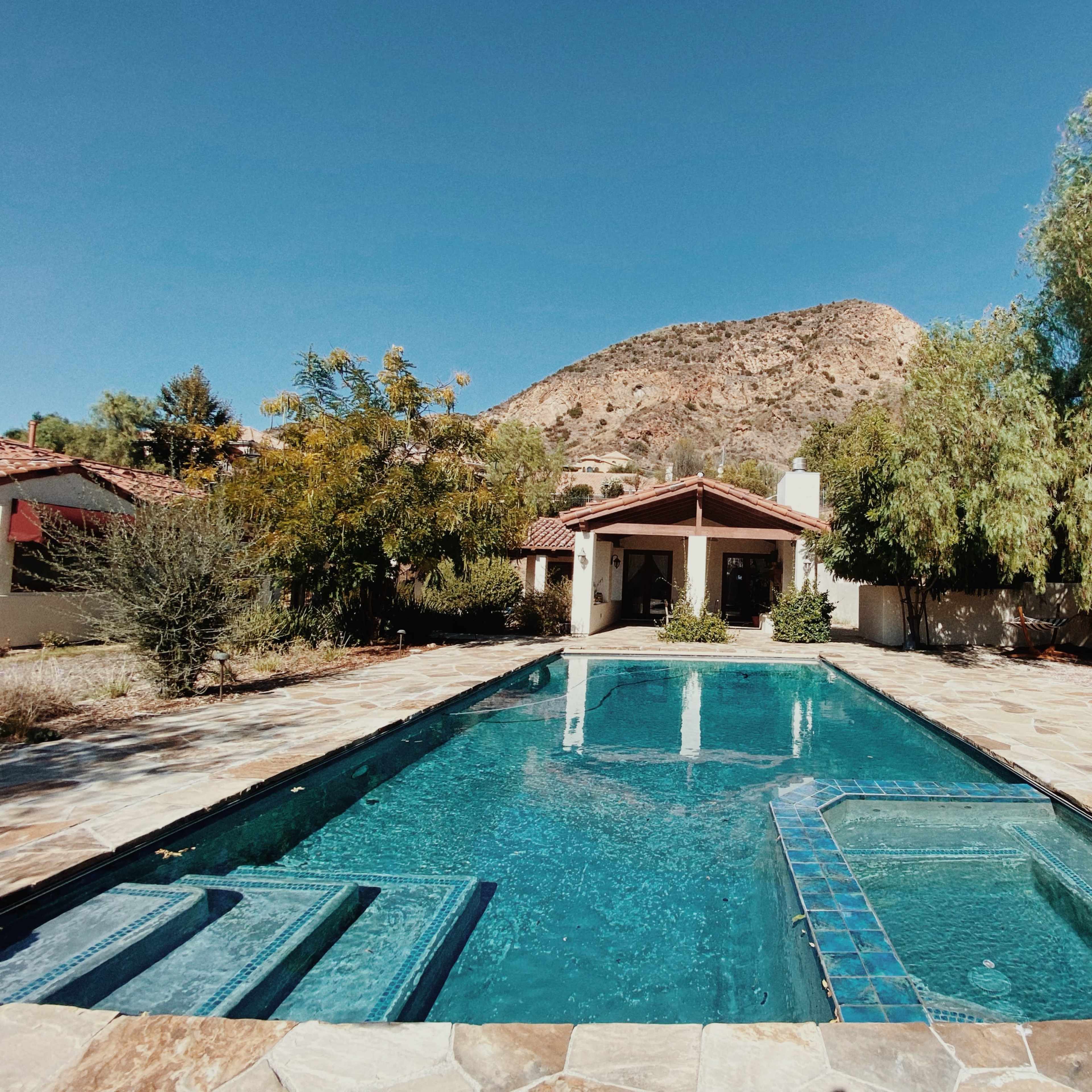 A rectangular swimming pool with a hot tub is surrounded by stone tiles and landscaping, set against a backdrop of a mountain on a clear day.
