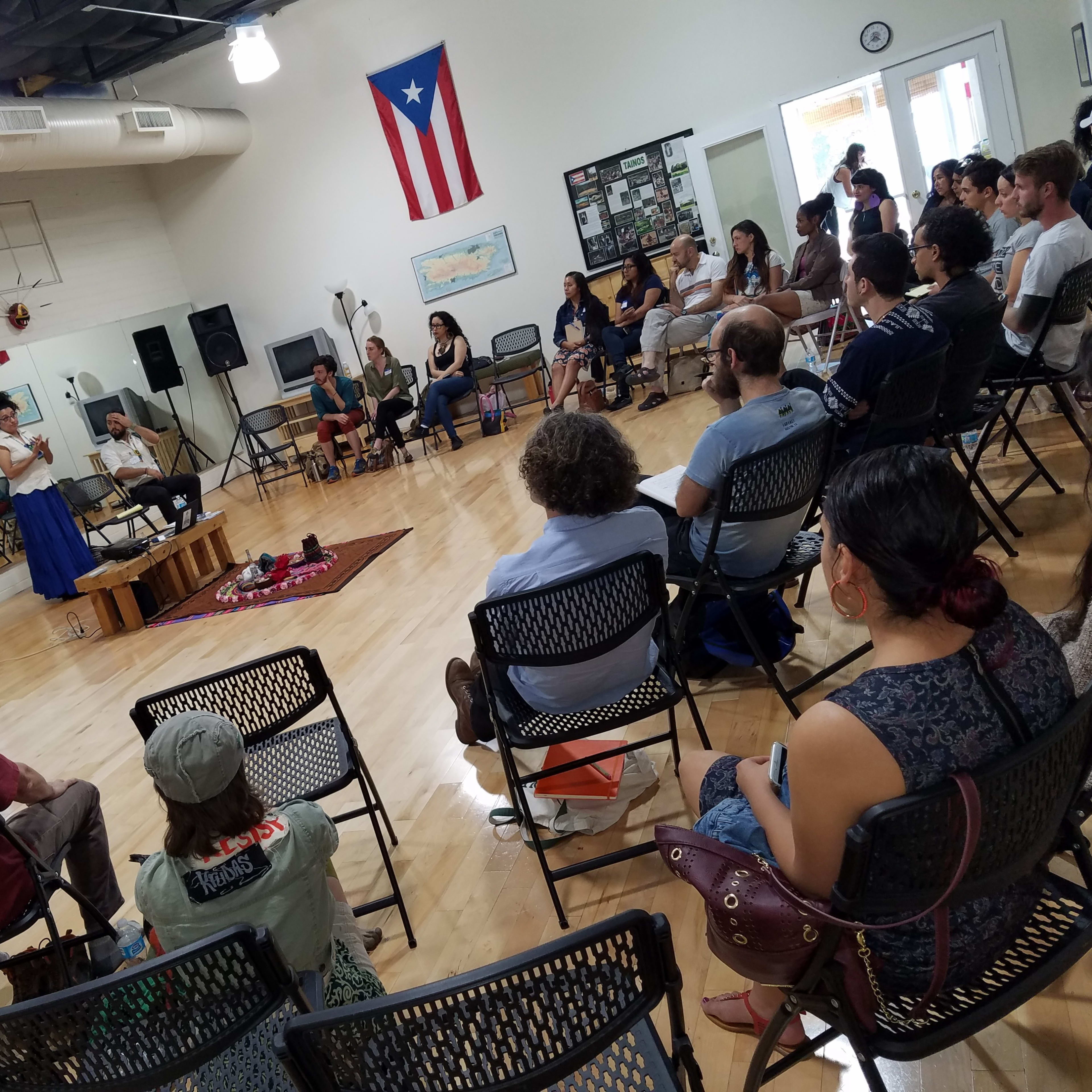 A speaker presents to an audience seated in a community center, with a projector screen displaying information in the background.