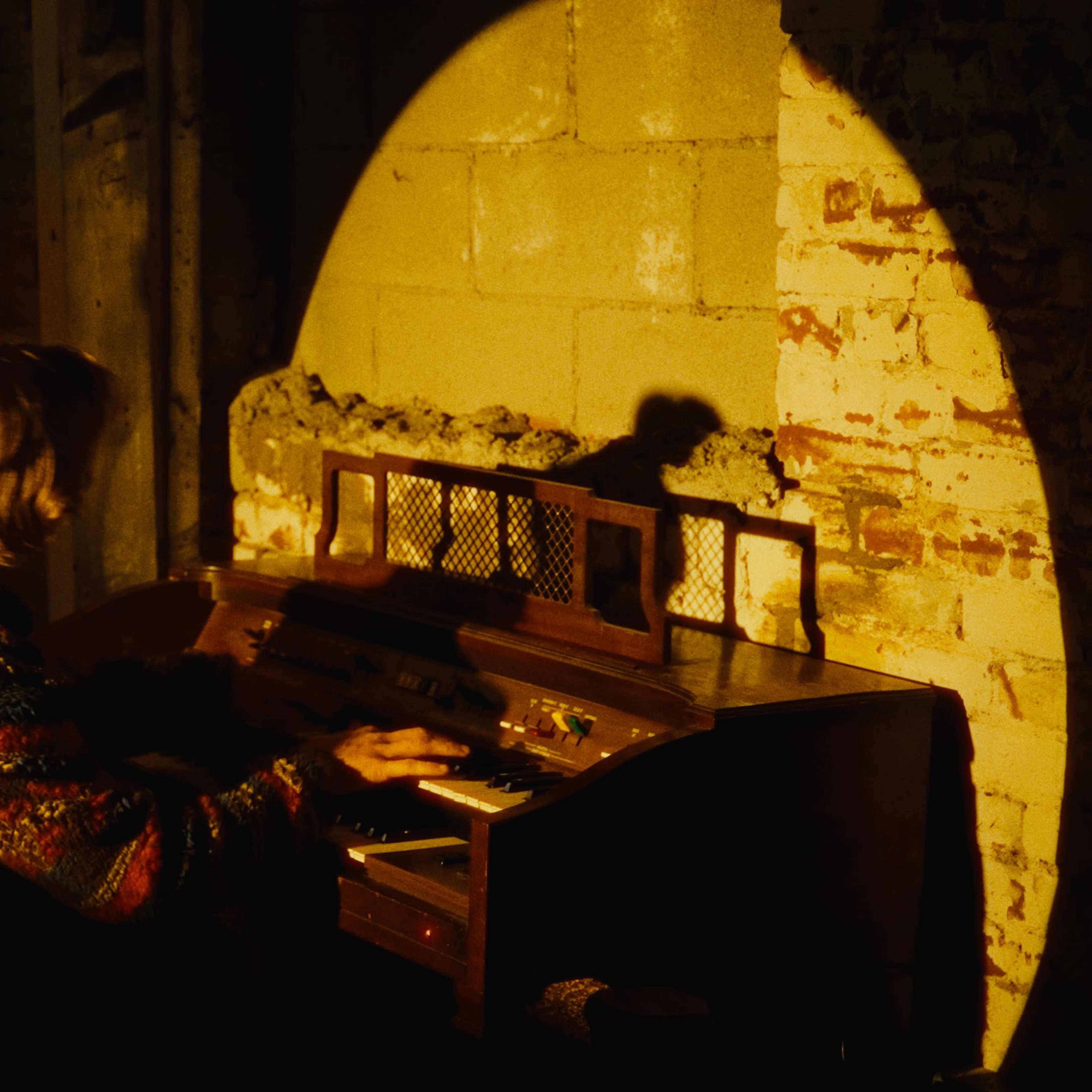 A person in a colorful sweater plays an organ in a dimly lit room with brick walls.