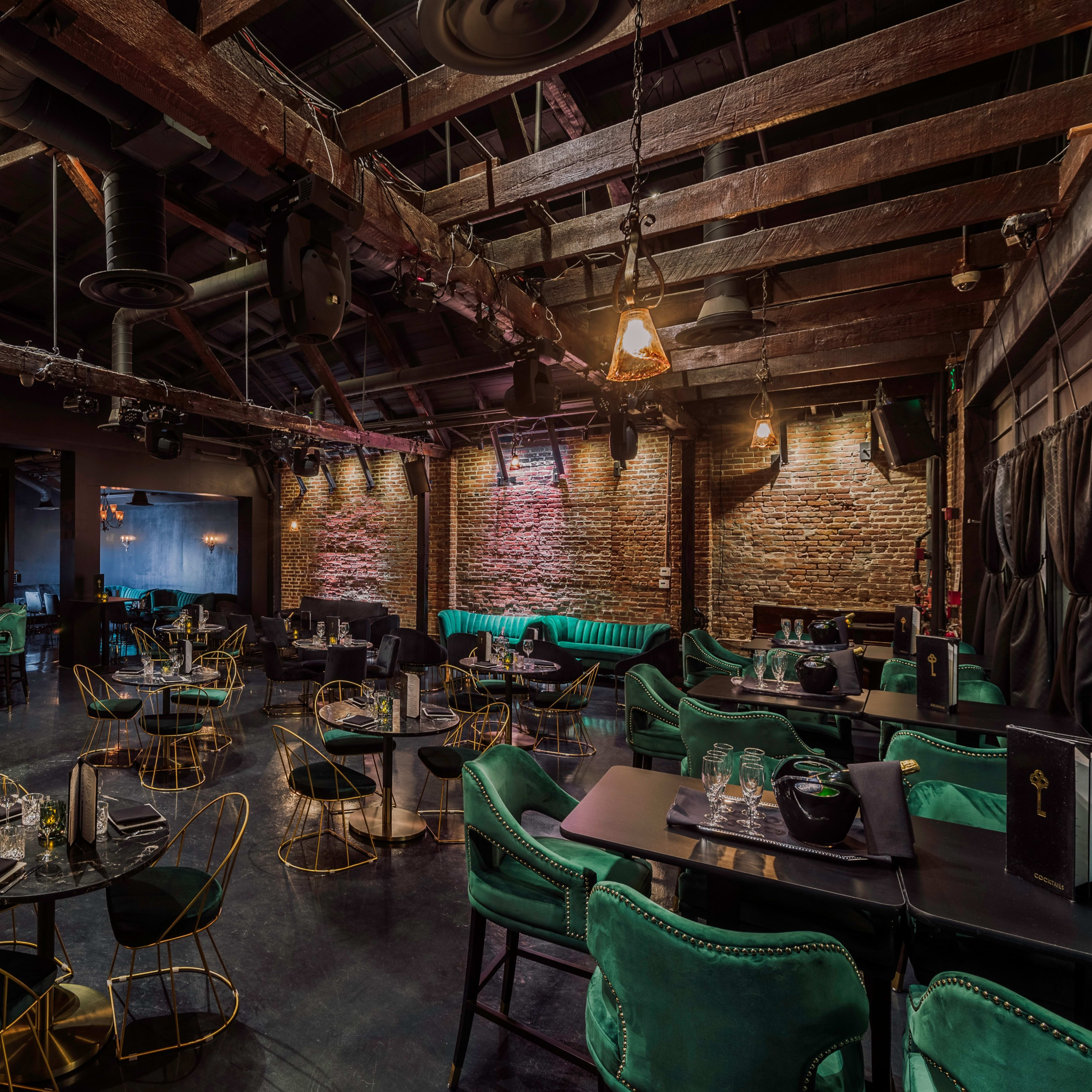 A dimly lit restaurant with wooden beams, exposed brick walls, and tables set with green upholstered chairs and black tablecloths.