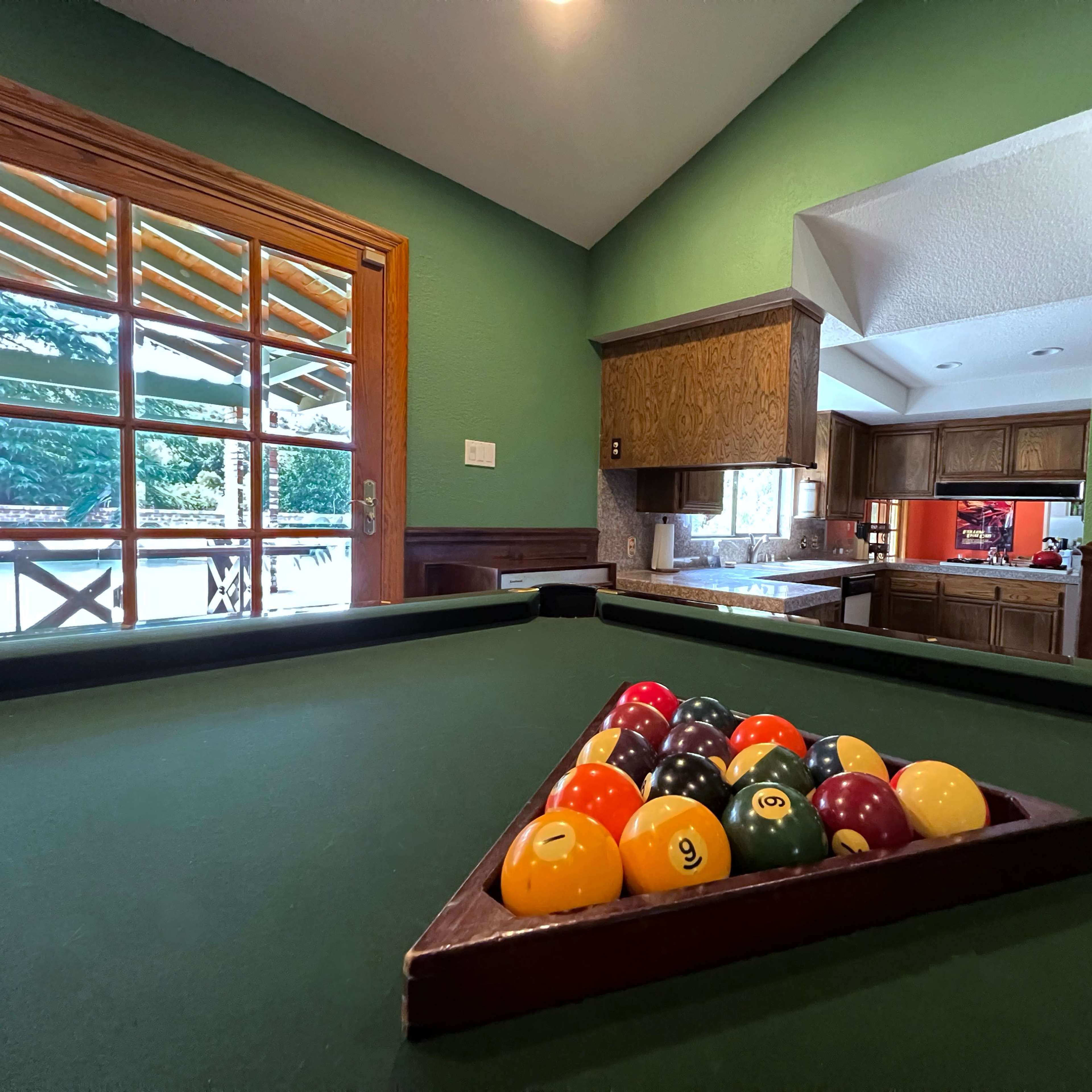 A billiards table with a triangular rack of colored pool balls is set in a room featuring green walls and large windows that overlook an outdoor space.