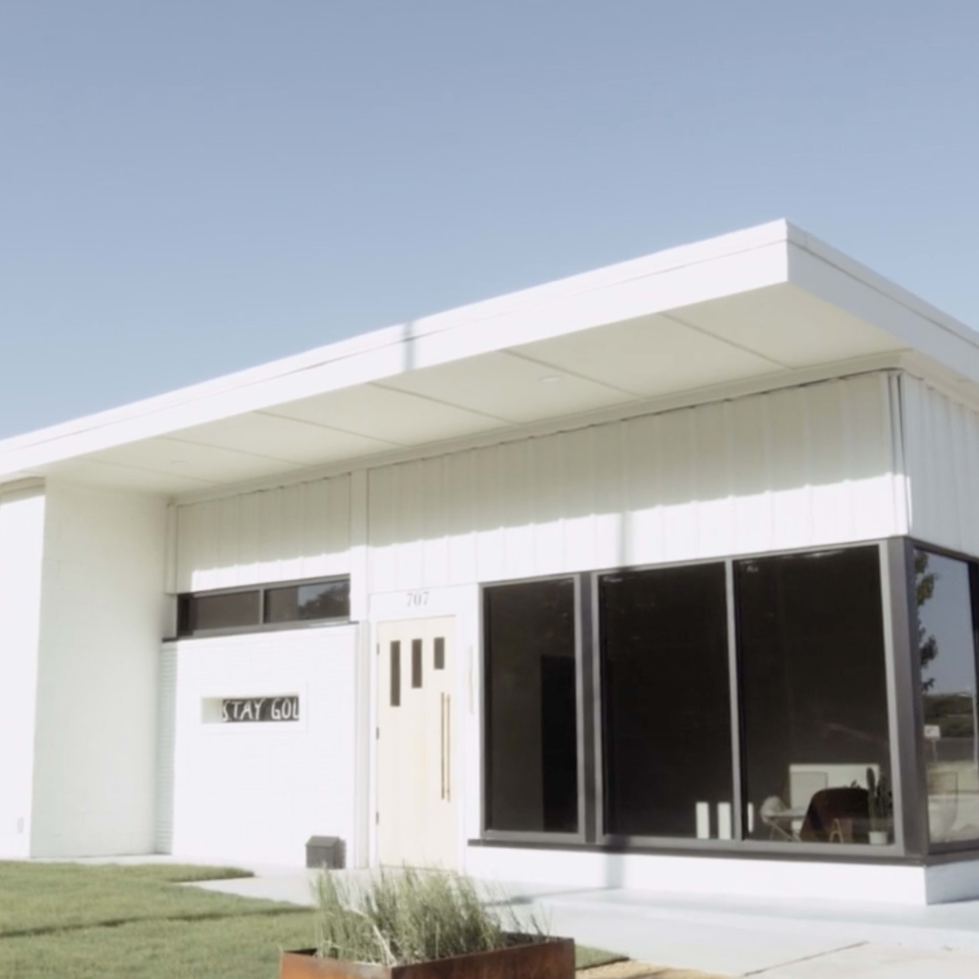 A modern single-story house with large glass windows and a flat roof, surrounded by a lawn and a gravel path.