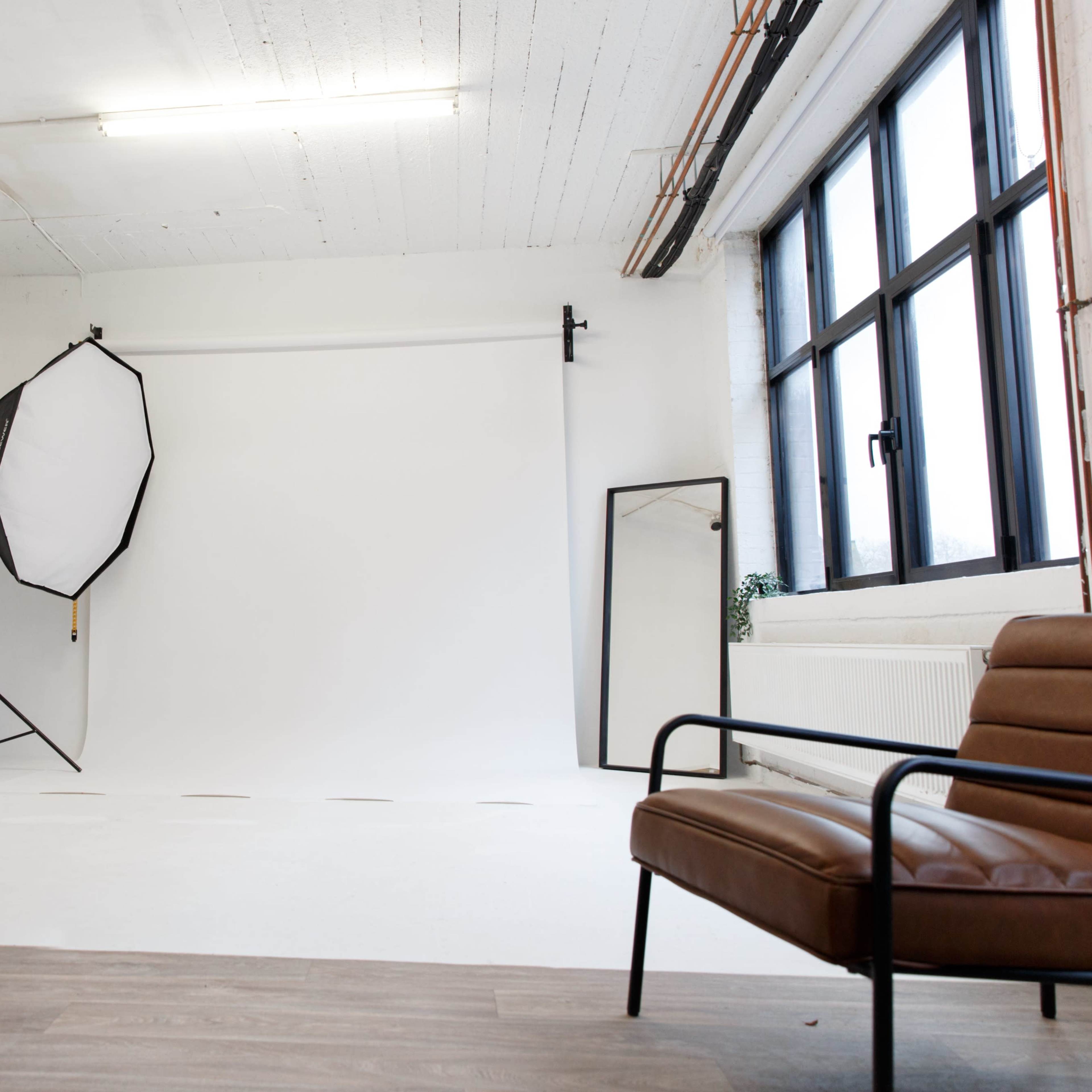 A minimalist studio space with a white backdrop, a large reflector setup on a stand, a mirror, a chair, and a table.