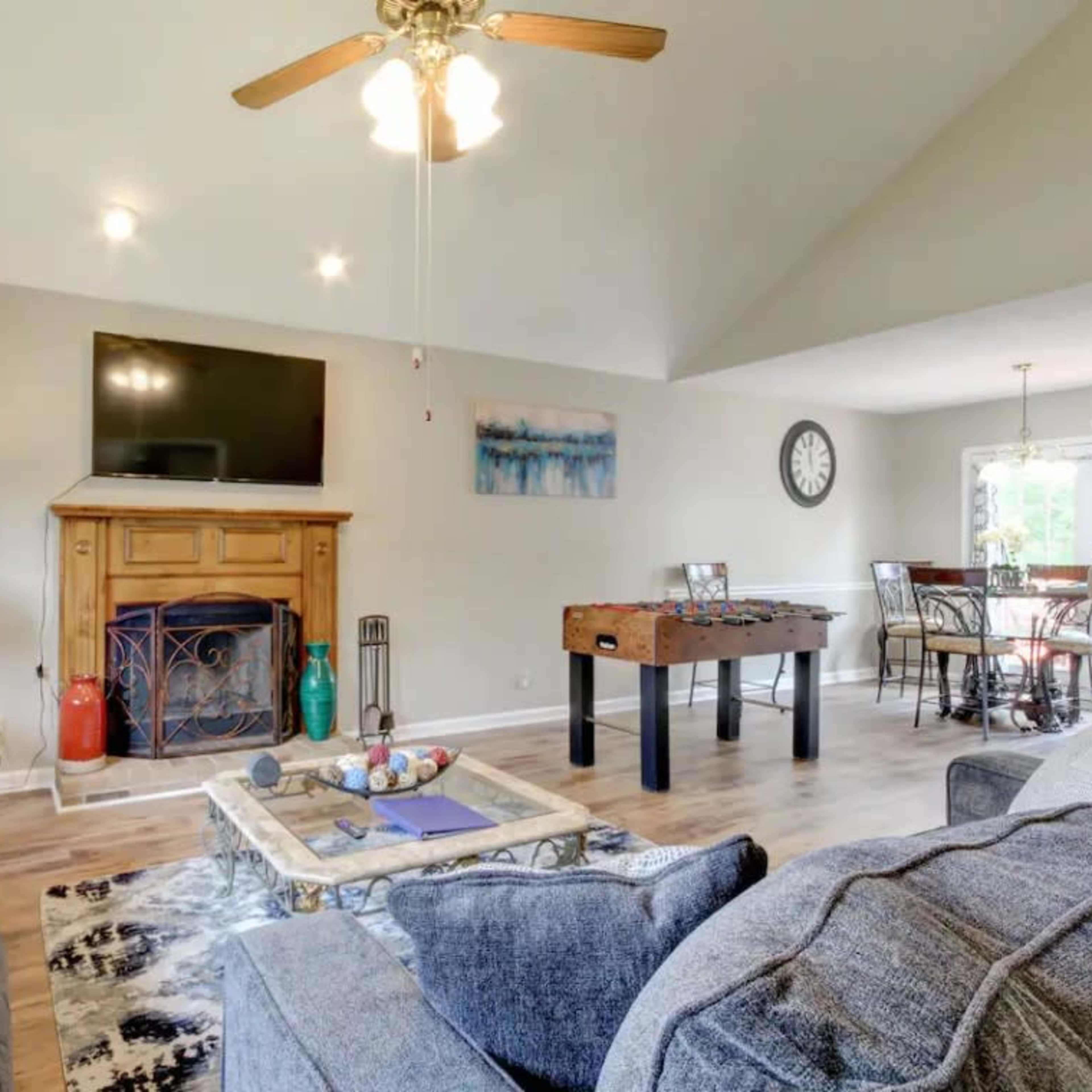 A living room with a sofa, a coffee table featuring decorative items, a fireplace beneath a wall-mounted television, and a foosball table nearby, with a dining area visible in the background.