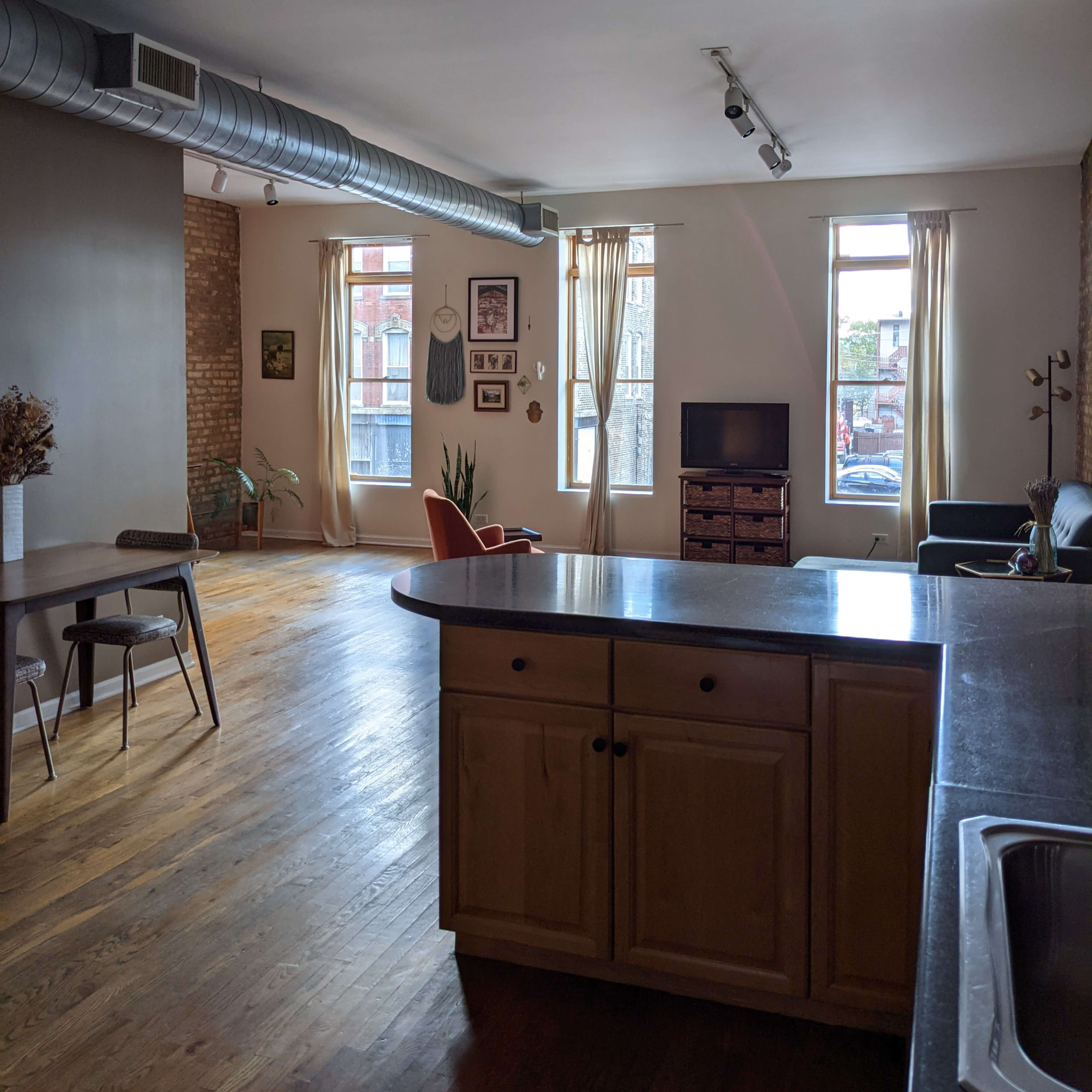 A spacious living area with a kitchen island, featuring wood flooring, large windows, and minimal furniture, including an orange chair and a television.