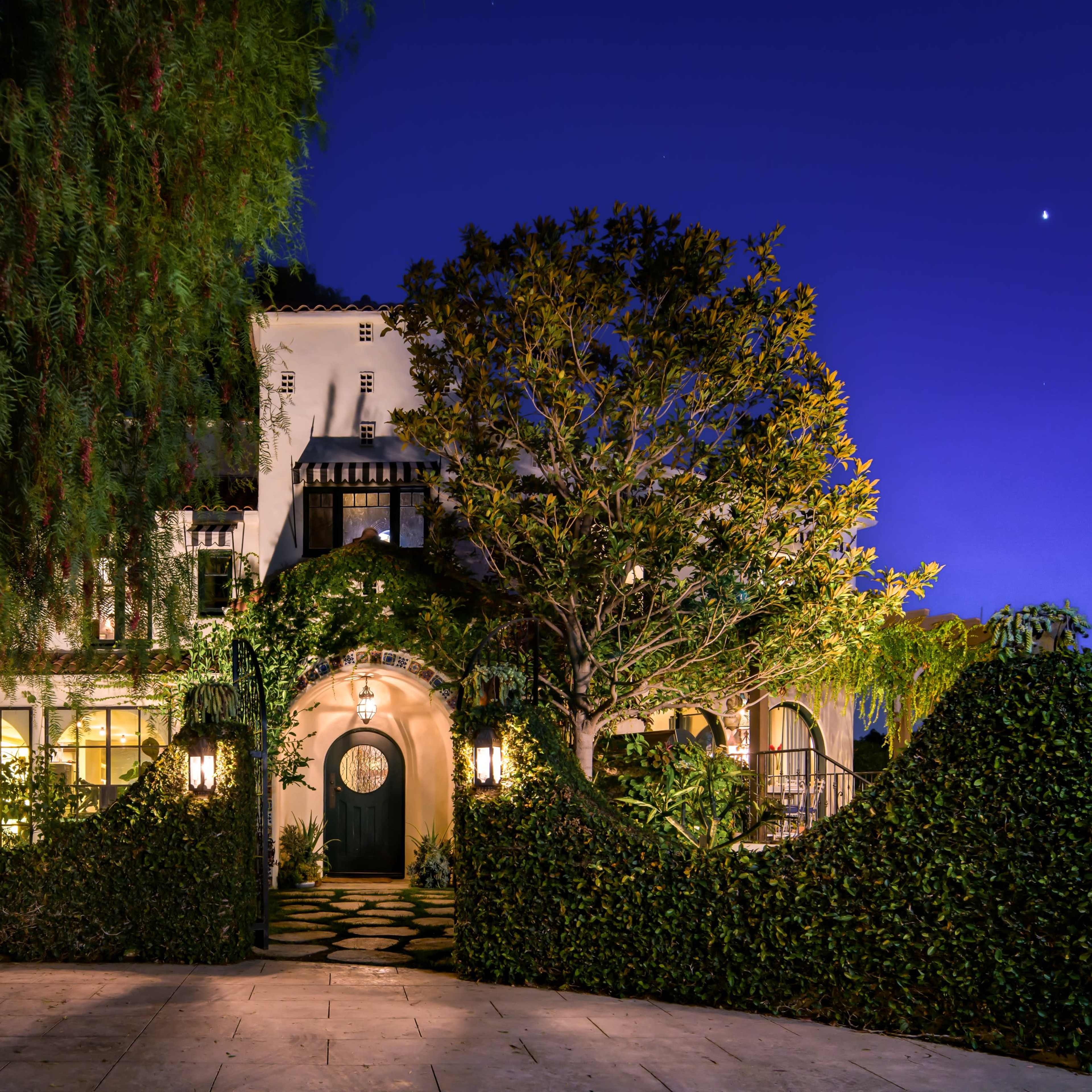 A two-story house covered in greenery and accented by outdoor lighting stands at dusk, with a round door and a stone pathway leading to it.