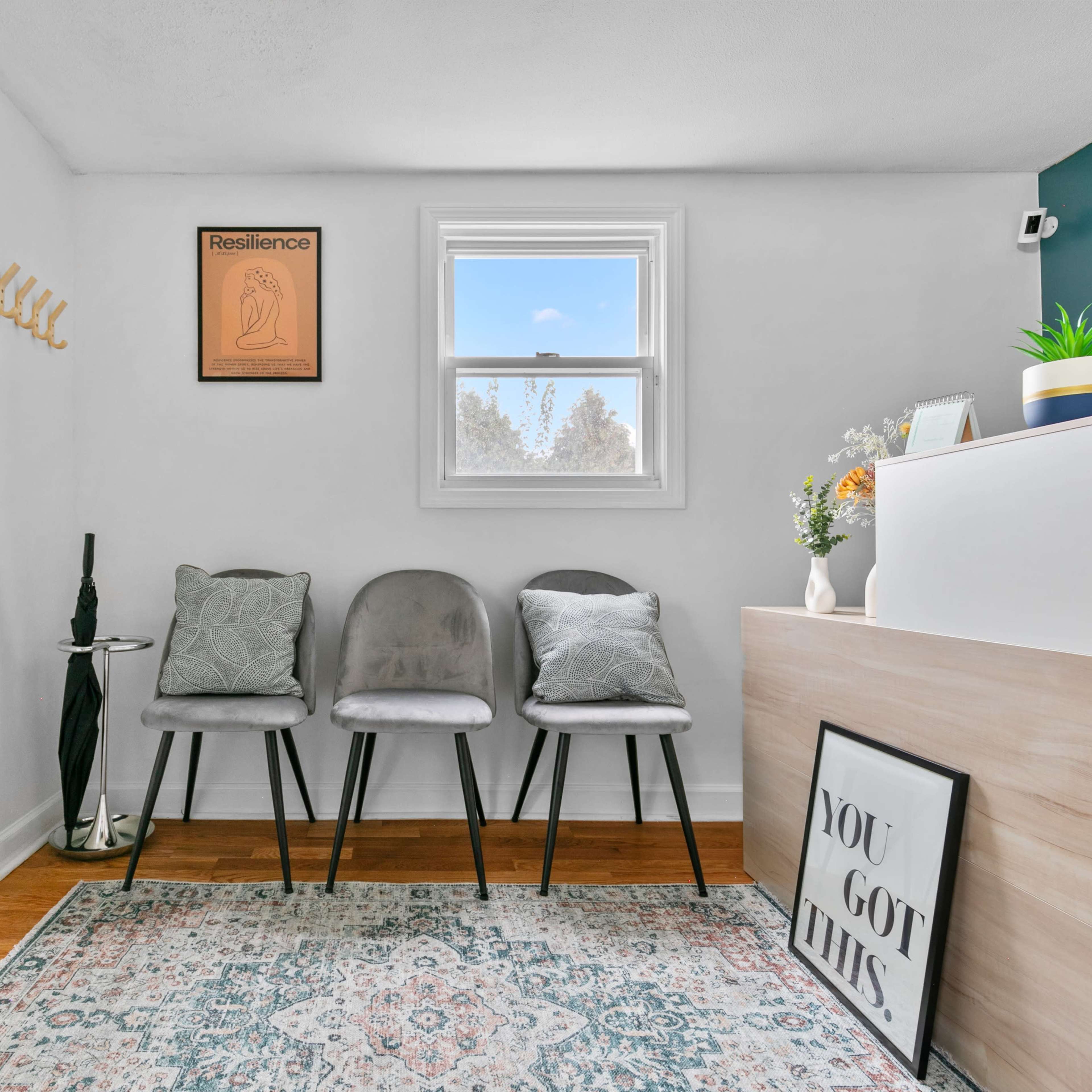 A small entryway features three gray chairs, a light wood reception desk, and a window allowing natural light to enter, with a decorative plant and artwork on the walls.