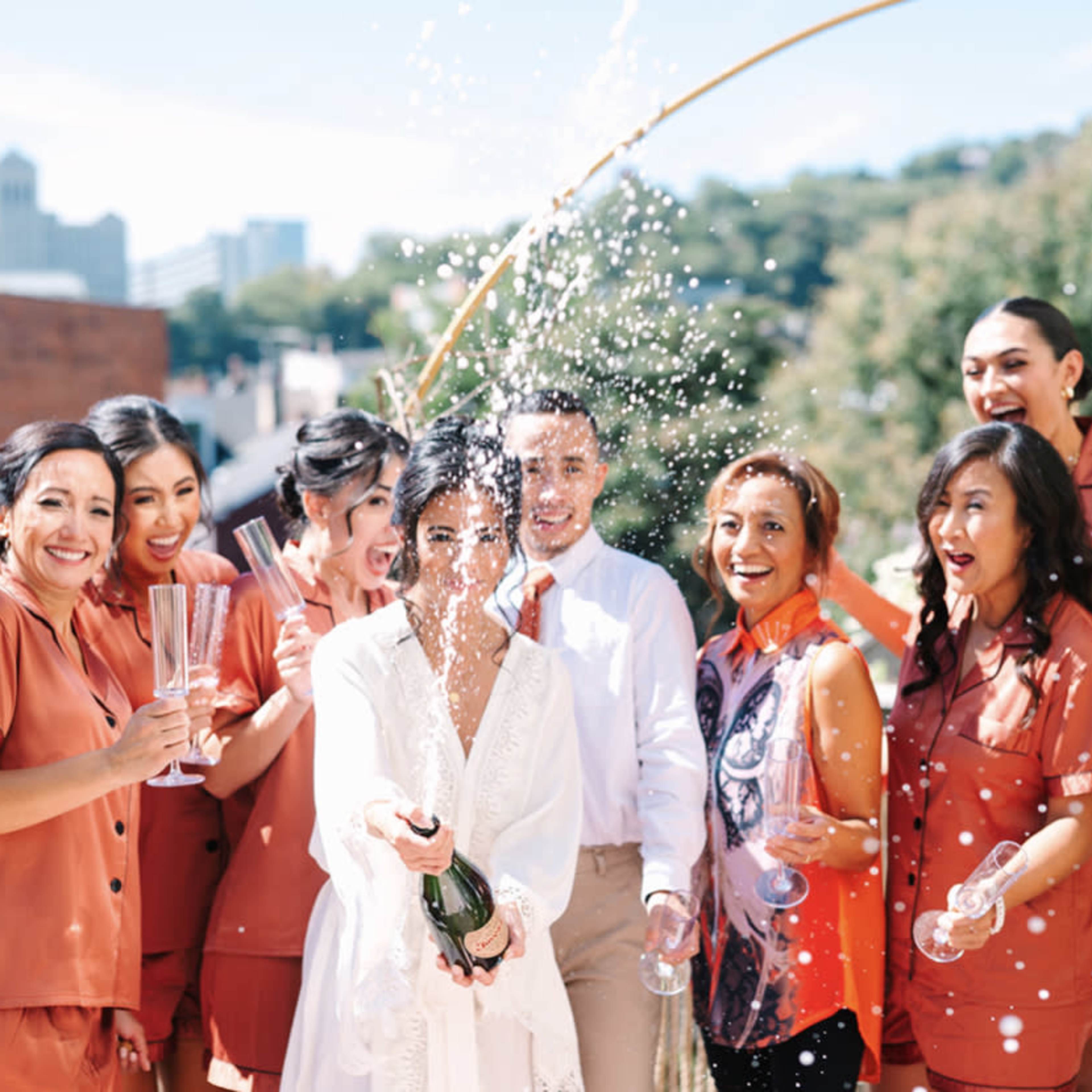 A group of women in matching orange outfits joyfully celebrates as one of them sprays champagne in an outdoor setting.