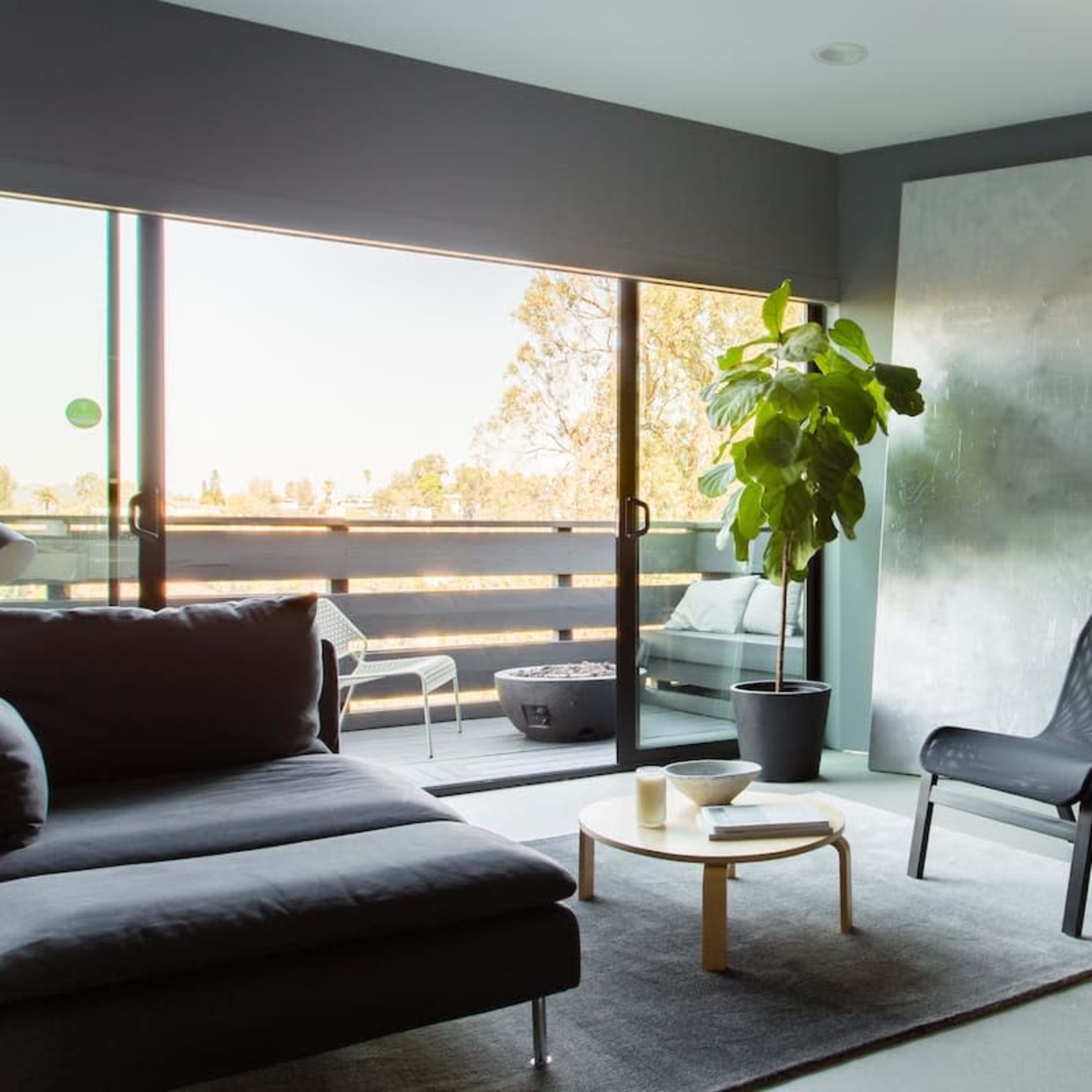 A modern living room features a gray sofa, a sleek chair, a round coffee table, and a large window overlooking an outdoor space with a plant beside it.