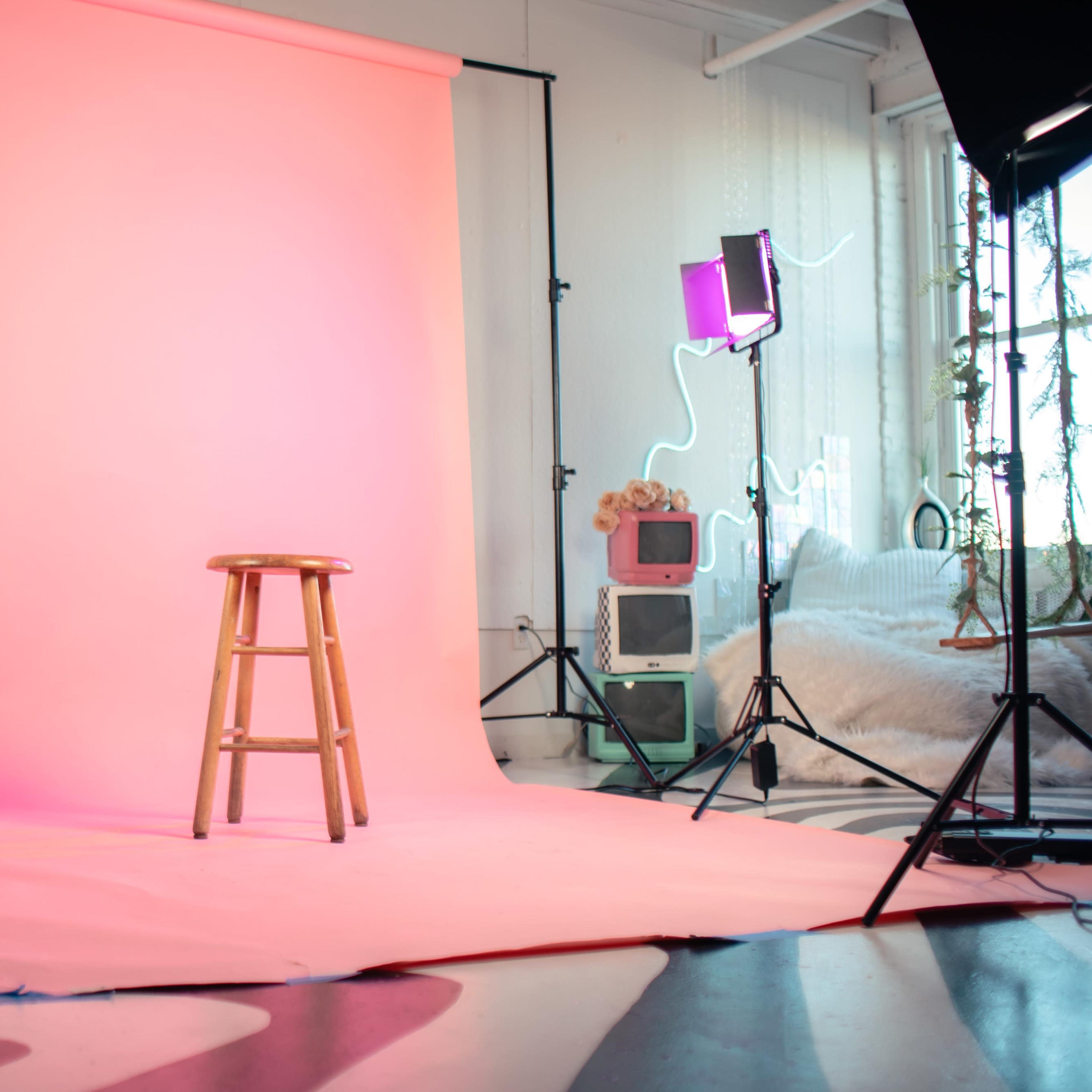 A photography studio setup featuring a pink backdrop, a wooden stool, and various lighting equipment arranged around the scene.
