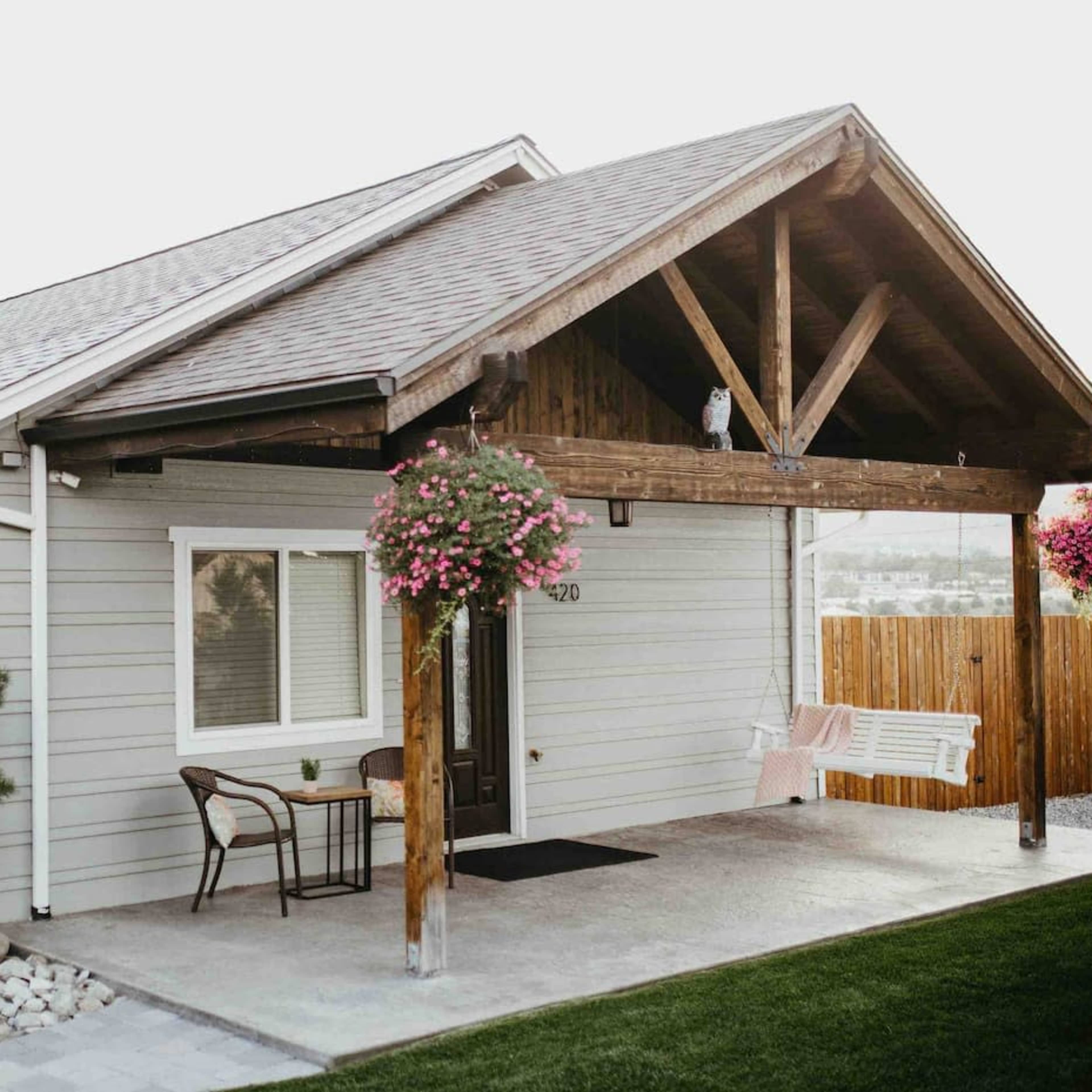 A single-story house with a gabled roof features a front porch supported by wooden beams, surrounded by landscaped greenery and hanging flower baskets.