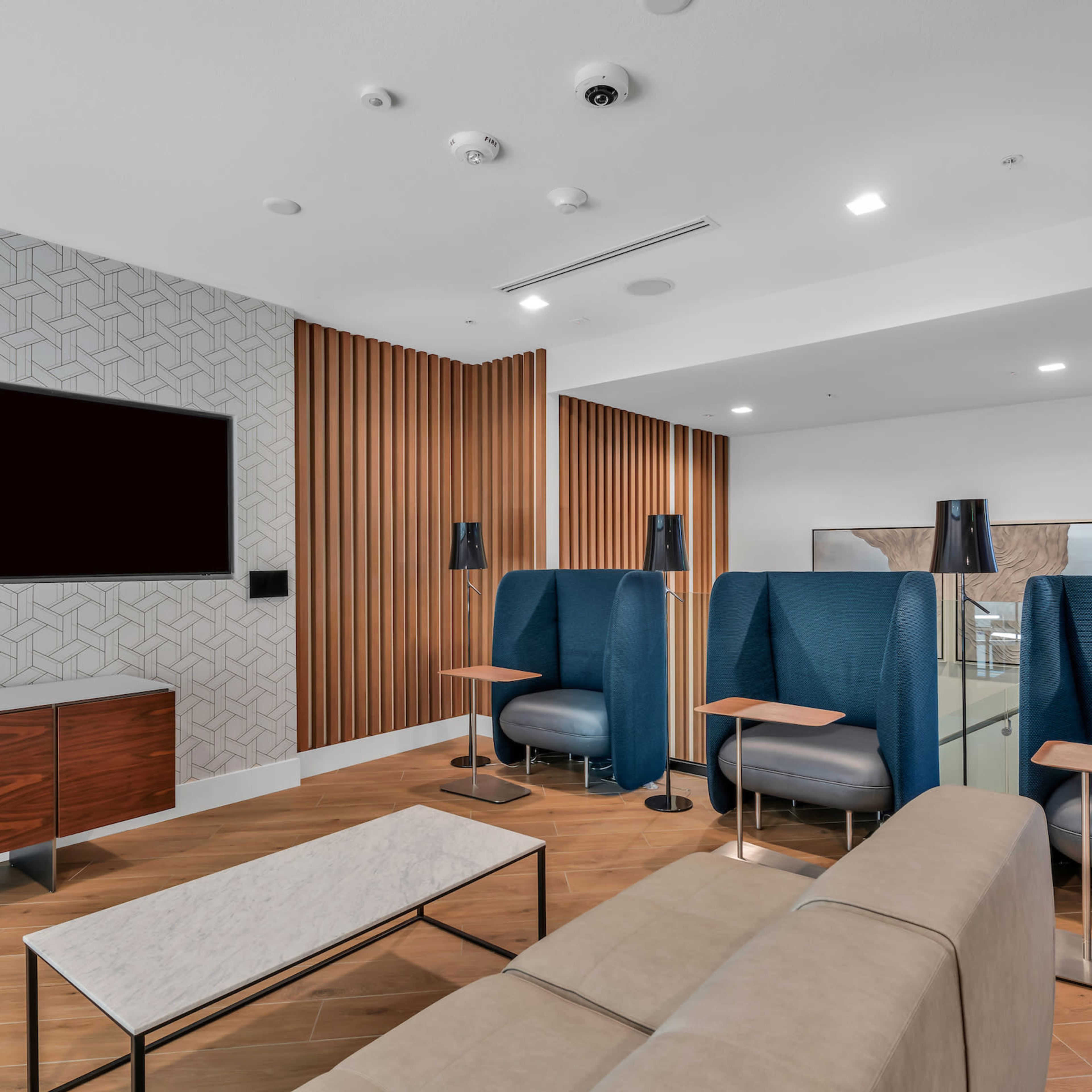 A modern lounge area featuring a beige couch, two blue wingback chairs, a coffee table, and a large wall-mounted television.