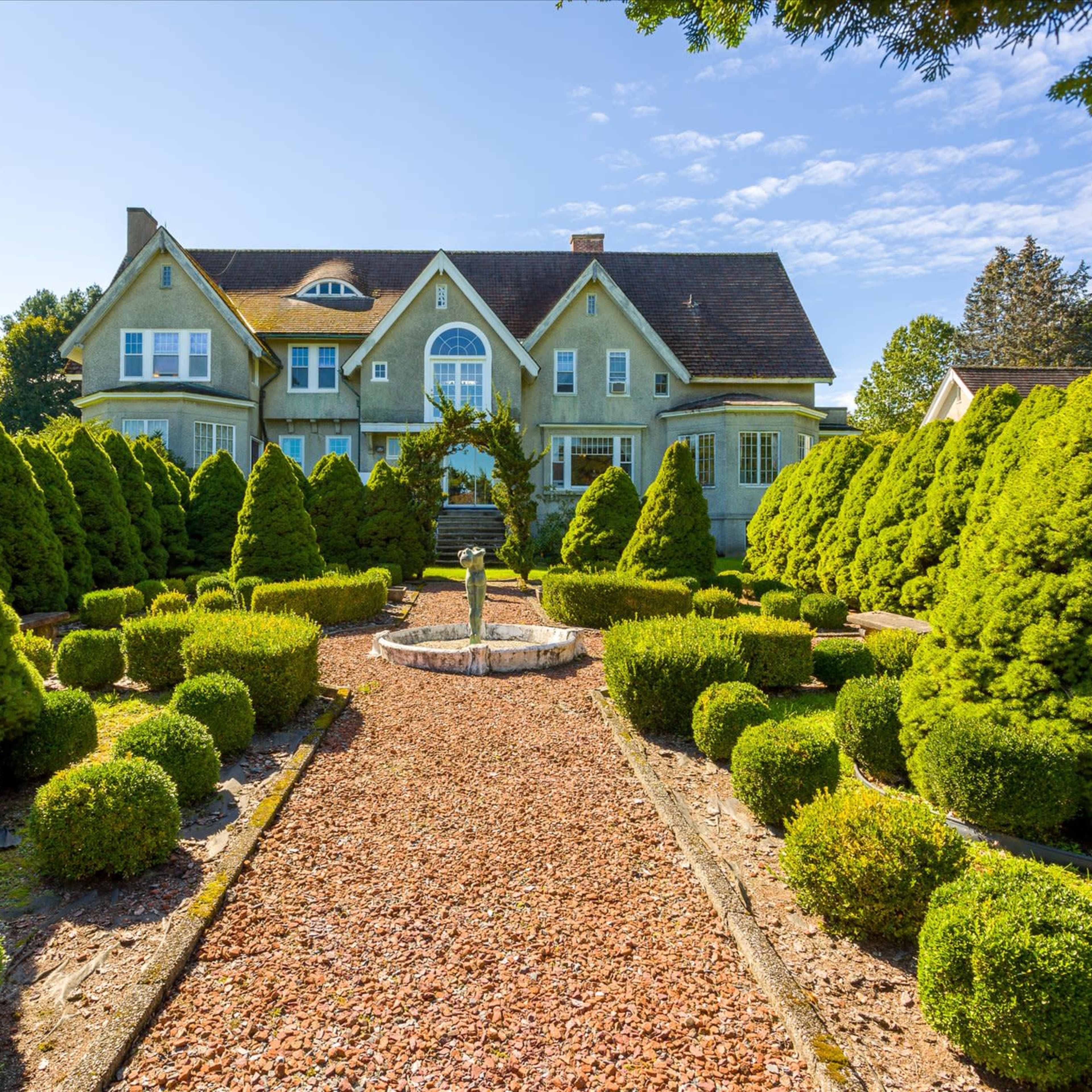 A large, elegant house surrounded by meticulously manicured hedges and a gravel path leading to a central fountain.