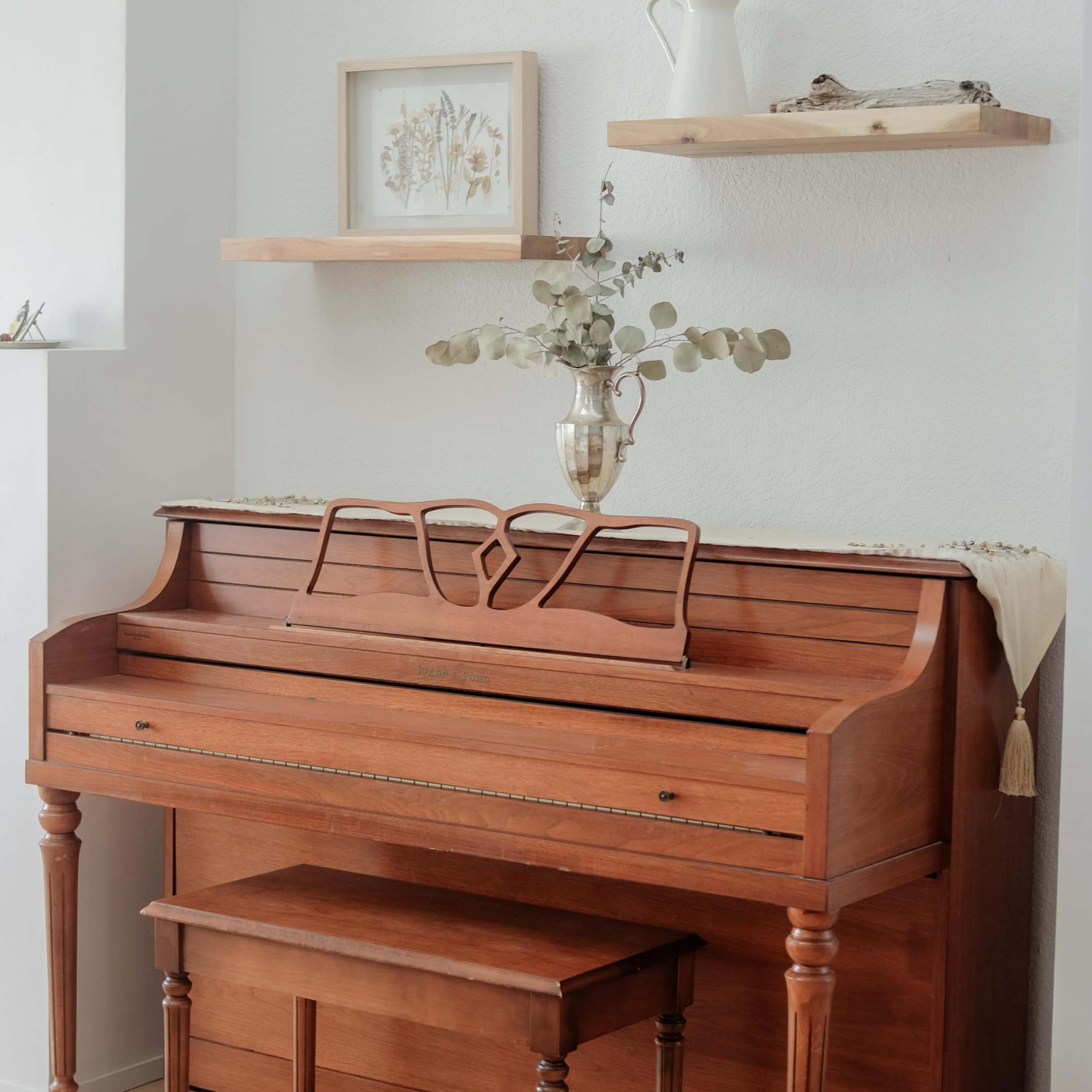 A wooden piano is positioned against a light-colored wall, accompanied by shelves displaying decorative items and a vase on top.